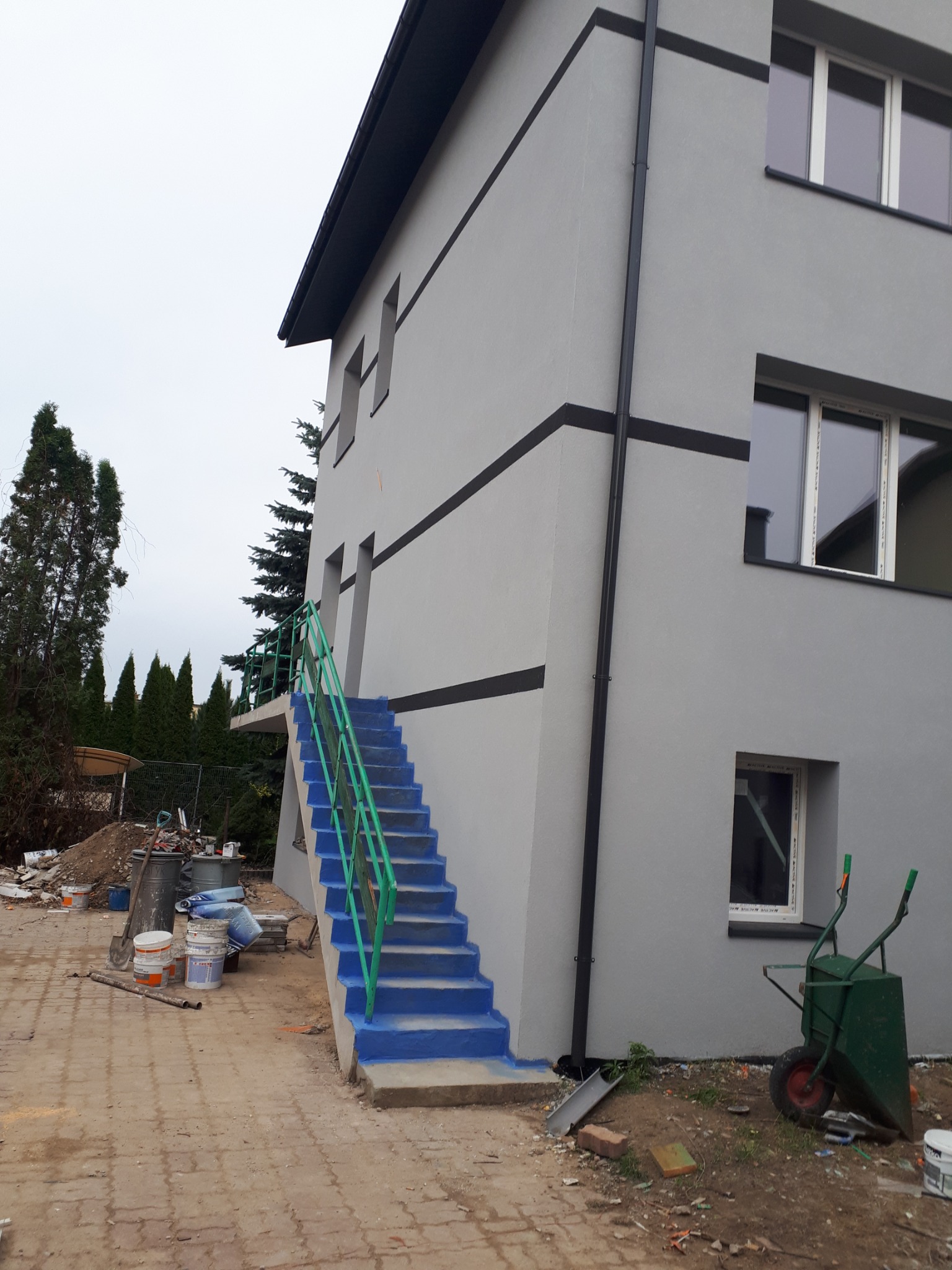 Szary budynek po renowacji elewacji z niebieskimi schodami i zieloną barierką, widoczne puszki po farbie i taczka.