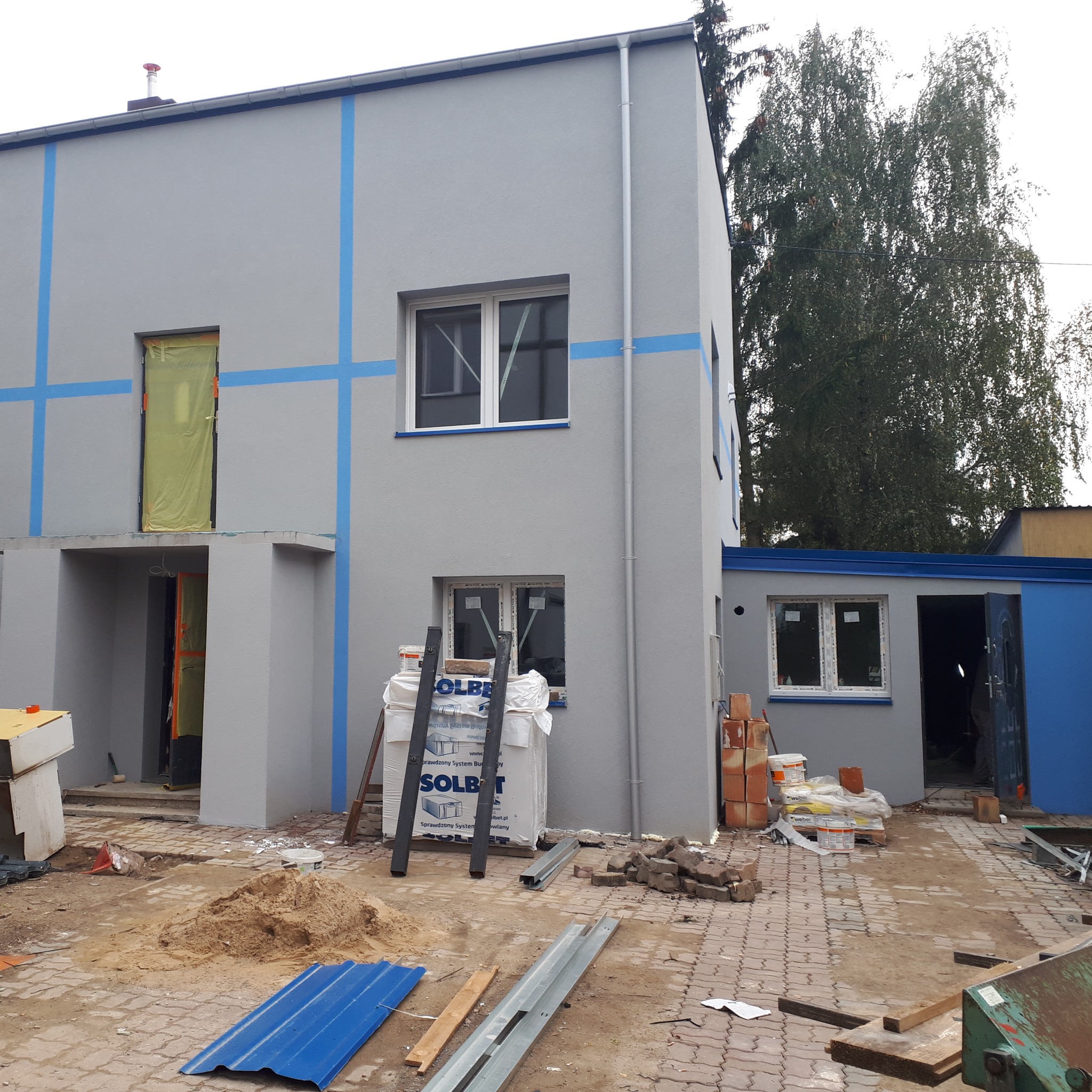 Dwukondygnacyjny budynek w trakcie prac elewacyjnych, szara fasada z niebieskimi akcentami, widoczne materiały budowlane, rusztowanie i teren budowy.