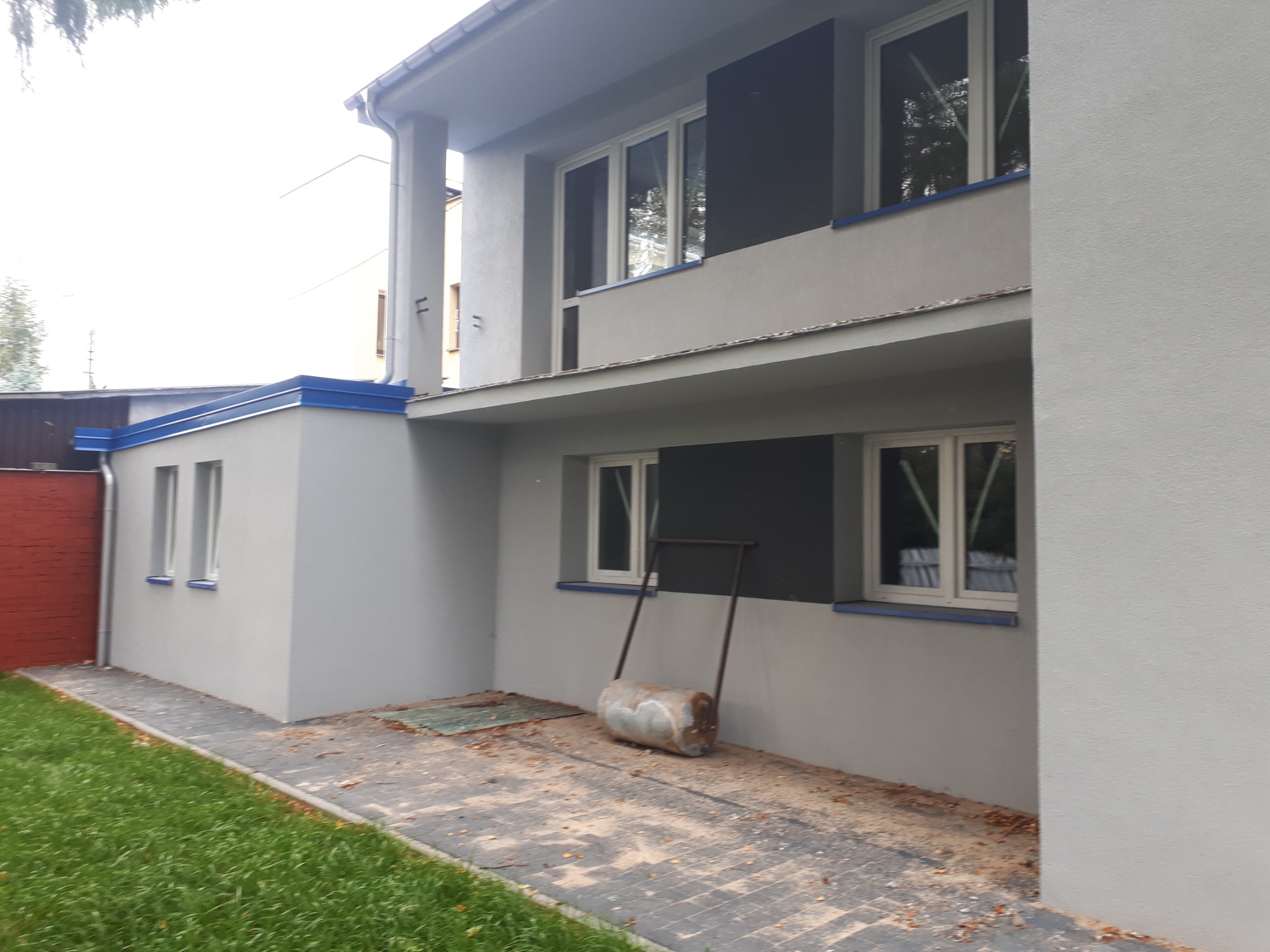 Odnowiona elewacja dwukondygnacyjnego domu z szarą fasadą, niebieskimi akcentami i metalowym wałkiem do trawy obok chodnika.