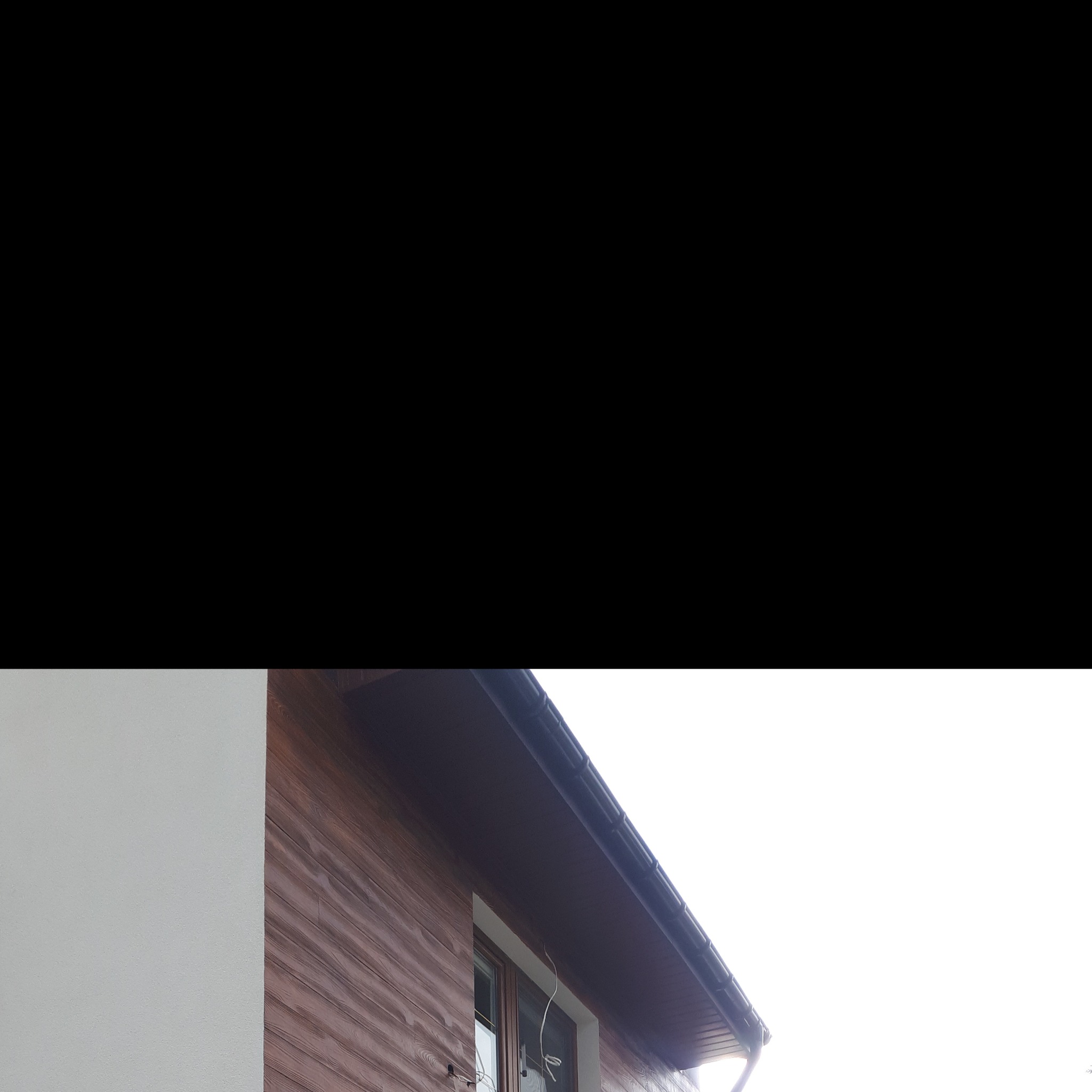 Fragment elewacji budynku z drewnianą okładziną i oknem, widoczne orynnowanie, ściana budynku pomalowana na biało.