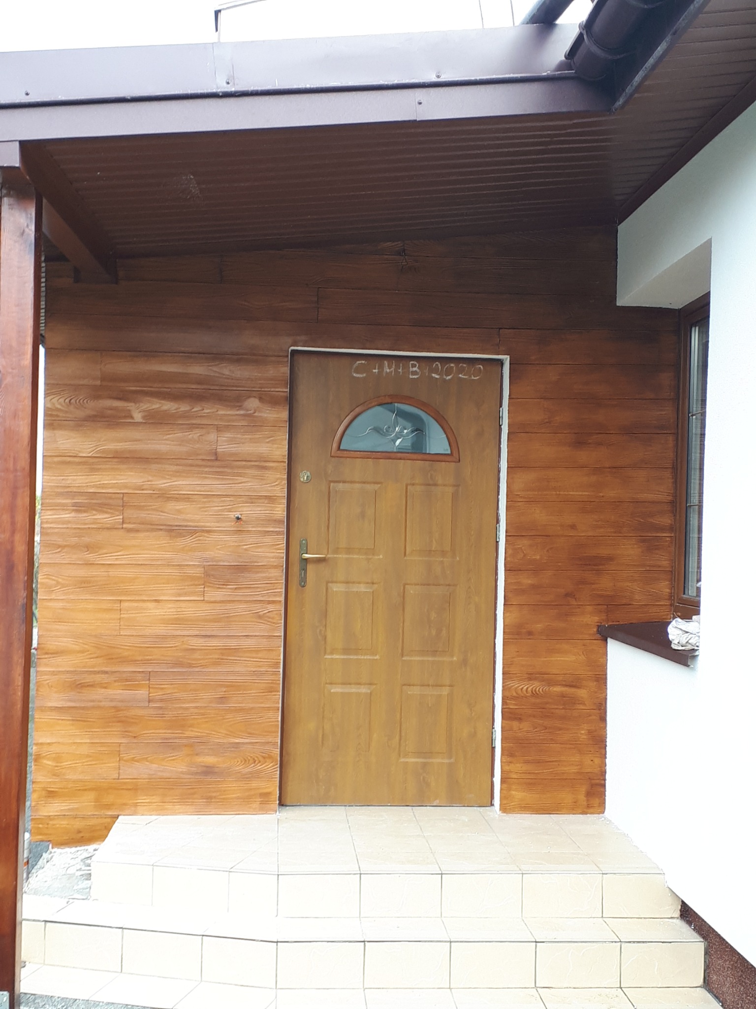 Wejście do domu z drewnopodobną elewacją, brązowymi drzwiami z szybką i białymi płytkami na schodach. Nad drzwiami widnieje napis 'C+M+B 2020'.