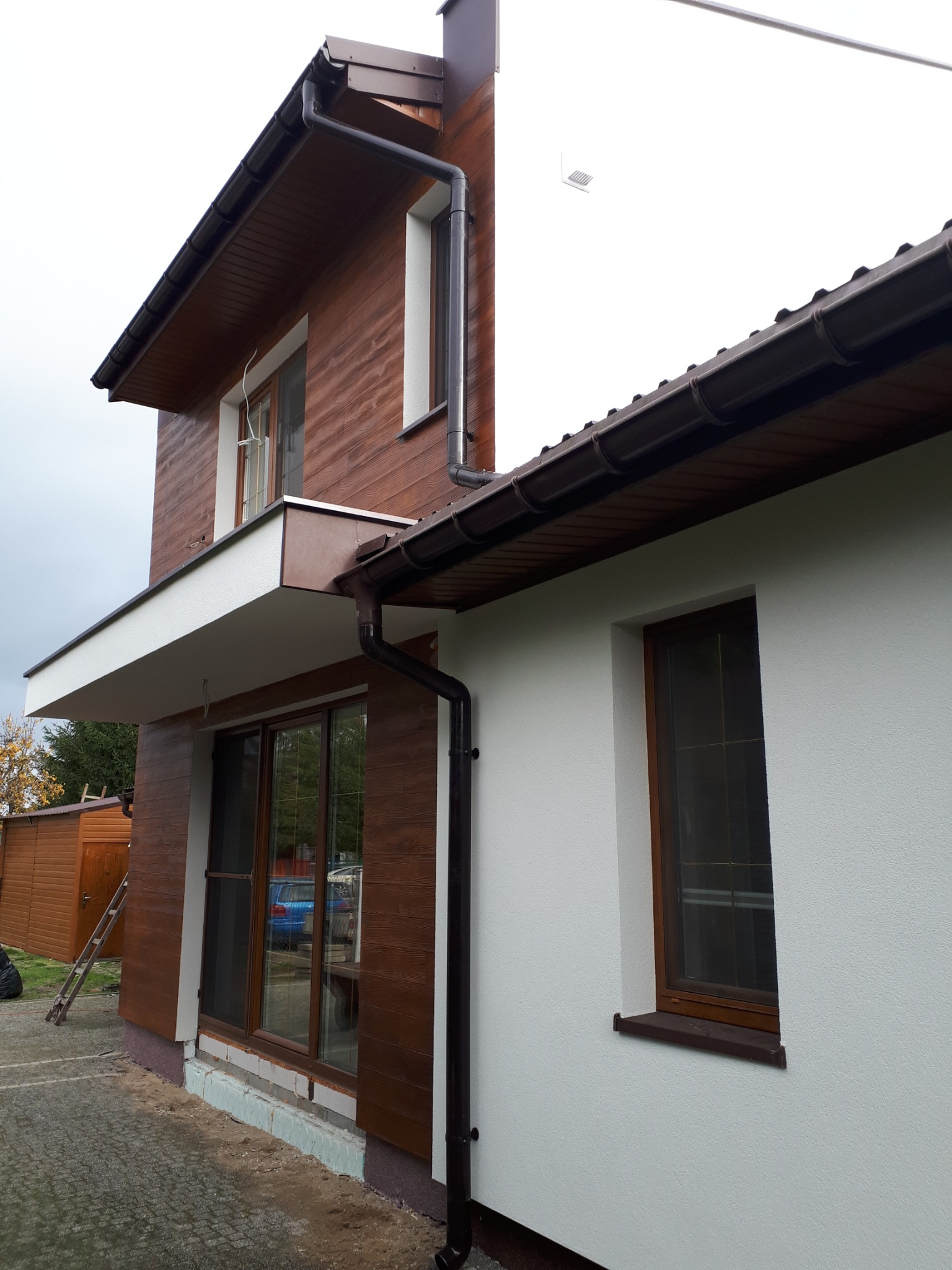 Dwukondygnacyjny dom z elewacją w kolorze białym i brązowym, z widocznymi rynnami i oknami z brązowymi ramami.