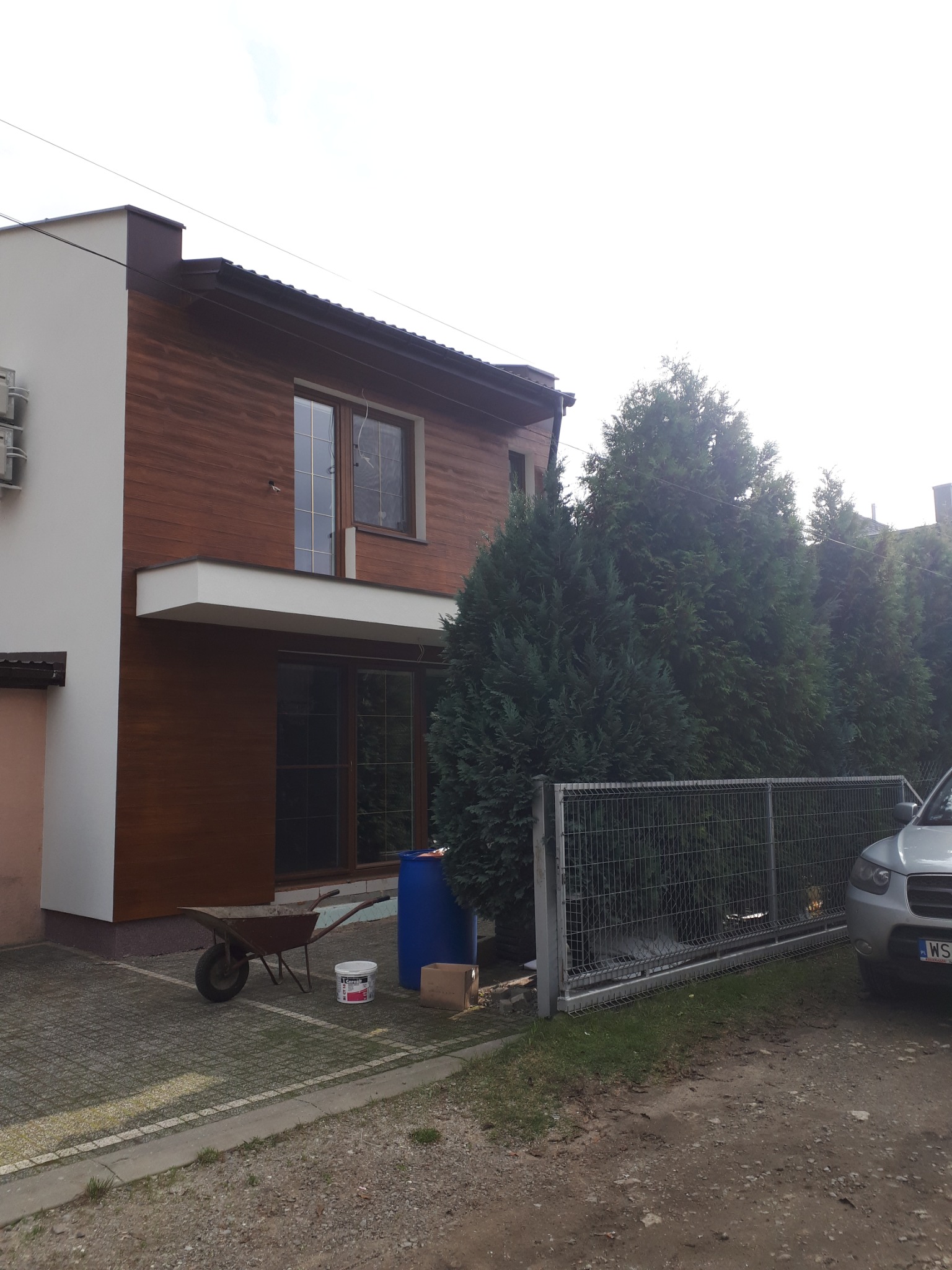 Dwupiętrowy dom z elewacją imitującą drewno, widok z zewnątrz, taczka i wiadro z farbą obok, sugerujące prace wykończeniowe.