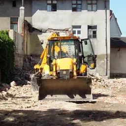 Żółta koparko-ładowarka stoi przed zrujnowanym budynkiem z gruzem, prawdopodobnie podczas prac rozbiórkowych.