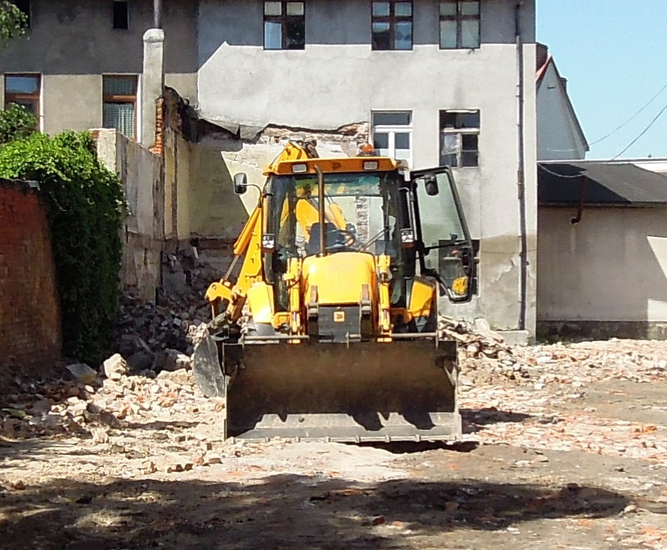 Żółta koparko-ładowarka stoi przed zrujnowanym budynkiem z gruzem, prawdopodobnie podczas prac rozbiórkowych.