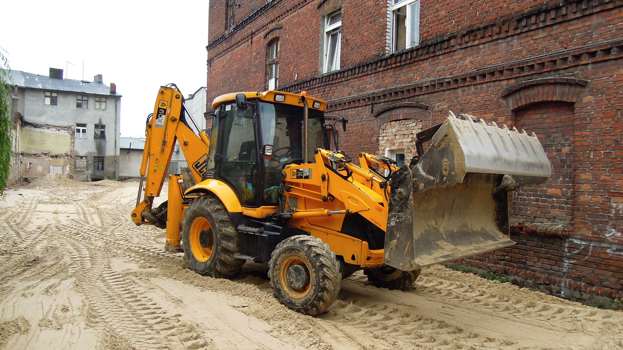Żółta koparko-ładowarka JCB stoi na piaszczystym terenie z widocznymi śladami opon, z przodu znajduje się duża łyżka, w tle ceglany budynek i budynek w trakcie remontu.