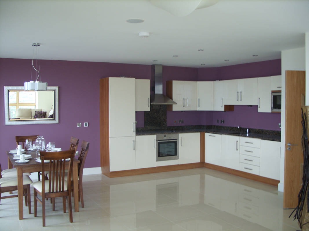 Jasna kuchnia z białymi szafkami i drewnianymi akcentami, fioletowa ściana, stół z zastawą stołową, odblaskowa podłoga z płytek.