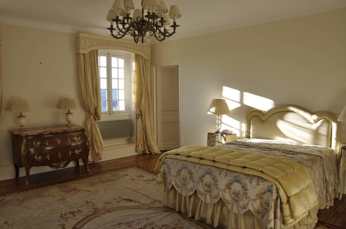 Elegancka sypialnia w stylu vintage z żyrandolem z brązu, komodą z marmurowym blatem i zasłonami w kolorze kremowym, oświetlona promieniami słońca wpadającymi przez okno.