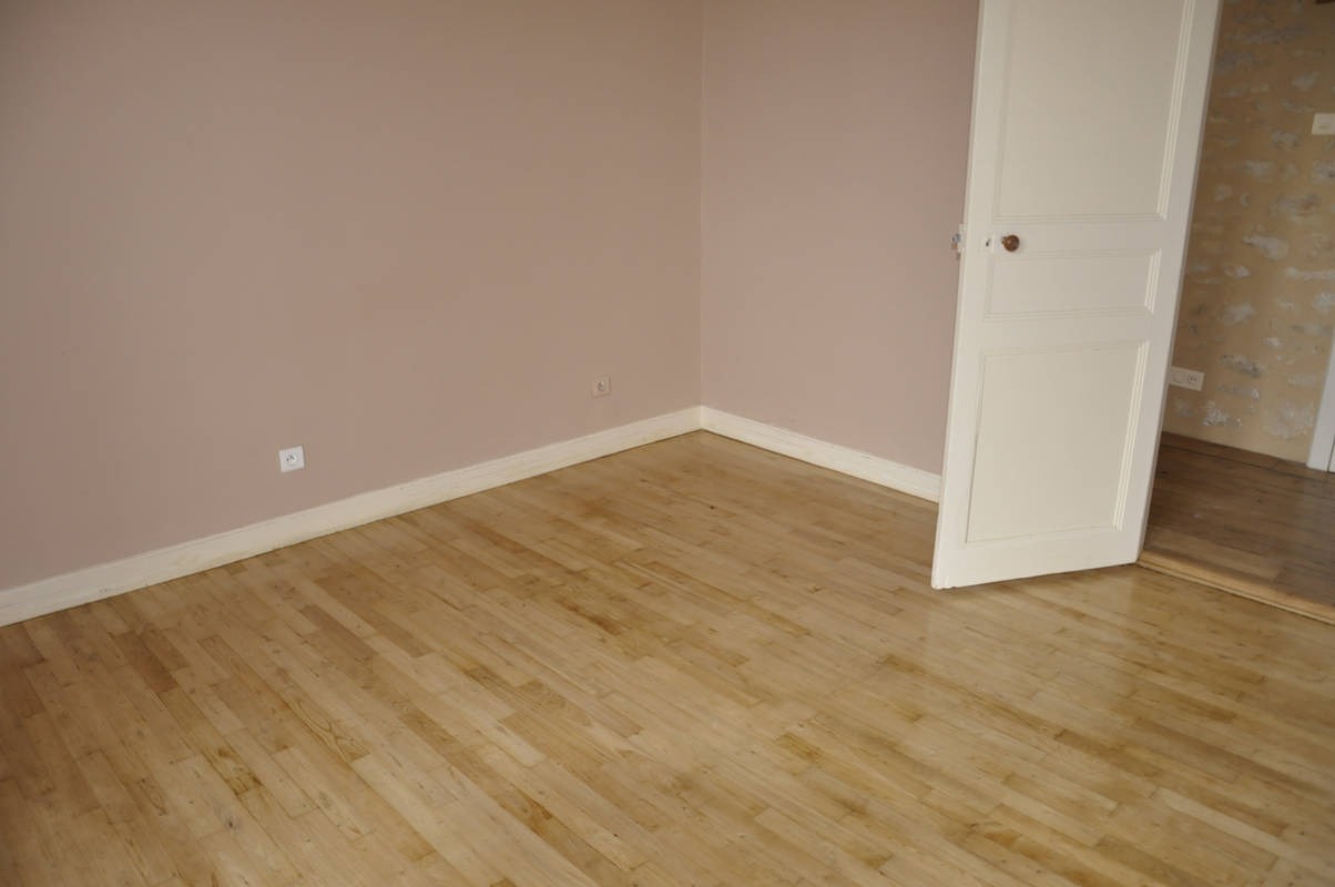 Pusty pokój z parkietem, ściany pomalowane na pudrowy róż, biała listwa przypodłogowa i otwarte białe drzwi z widokiem na kamienną ścianę w sąsiednim pomieszczeniu.