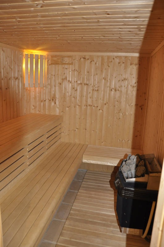 Wnętrze sauny z boazerią, drewnianymi ławkami i piecem z kamieniami, oświetlone ciepłym światłem.