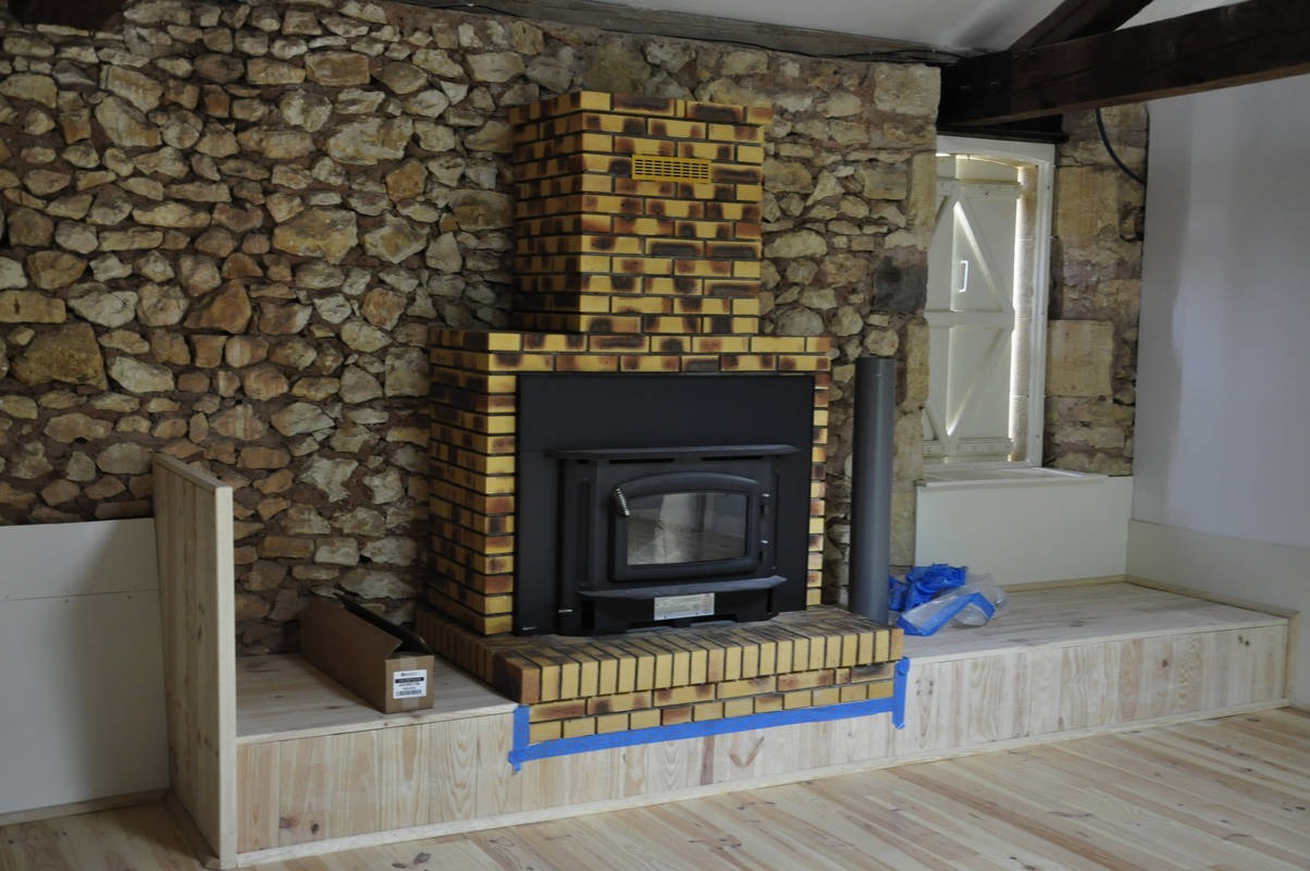 Wnętrze pokoju z kamienną ścianą i kominkiem z żółtej cegły, widoczny etap montażu obudowy kominka z drewnianych paneli, zabezpieczonych niebieską taśmą malarską.