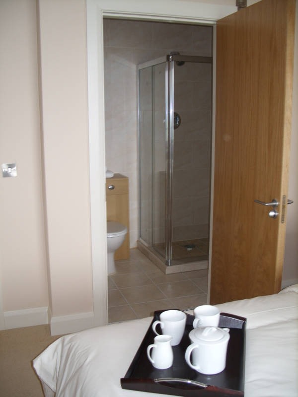 Widok z sypialni na łazienkę z kabiną prysznicową i toaletą, na pierwszym planie taca z białymi filiżankami i dzbankiem na łóżku.