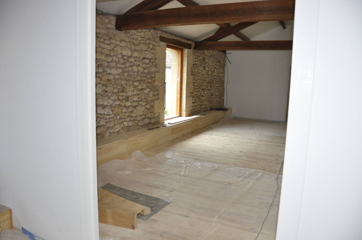 Wnętrze w trakcie prac wykończeniowych: kamienna ściana, drewniane belki stropowe, podłoga zabezpieczona folią malarską, widok zza odnowionych, białych drzwi.