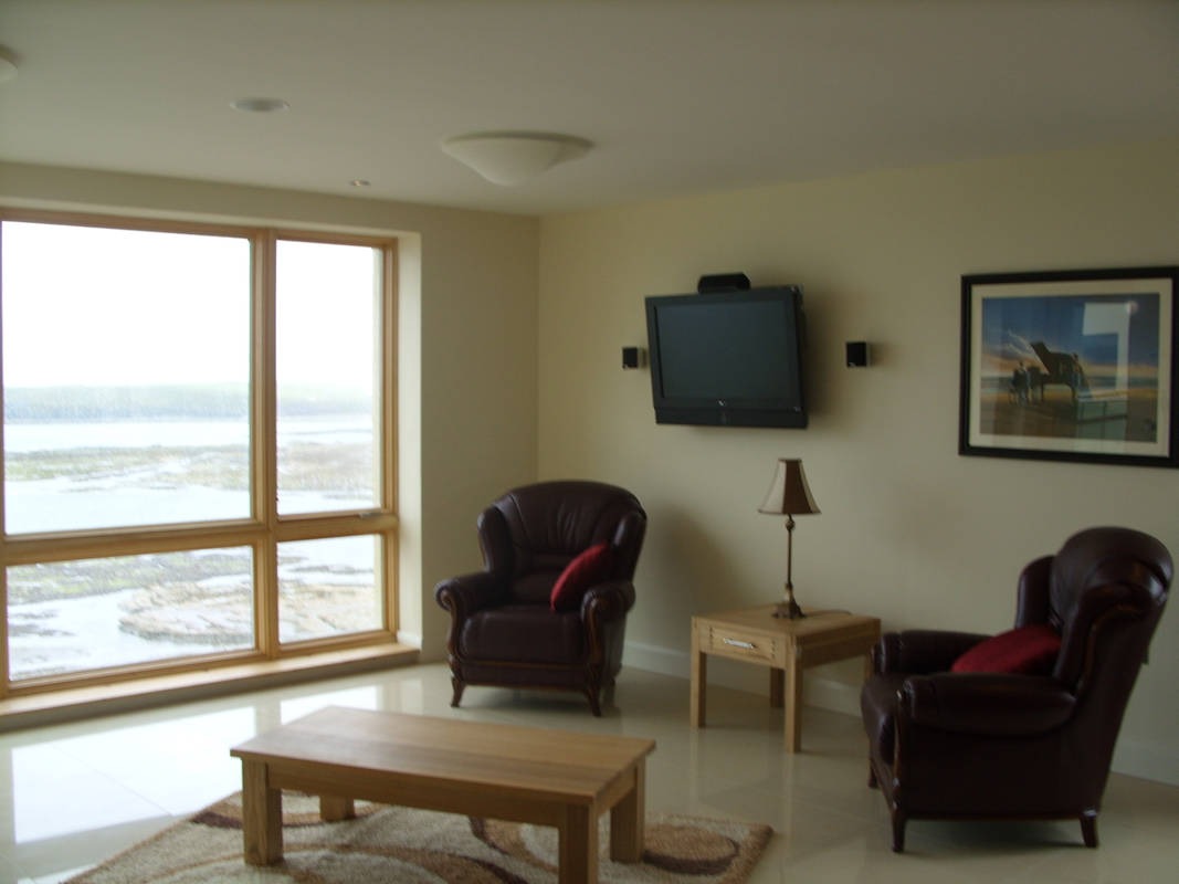 Jasne wnętrze salonu z dużym oknem z widokiem na morze, dwoma skórzanymi fotelami, drewnianym stolikiem kawowym na wzorzystym dywanie, lampą stołową i telewizorem na ścianie, całość w neutralnej...