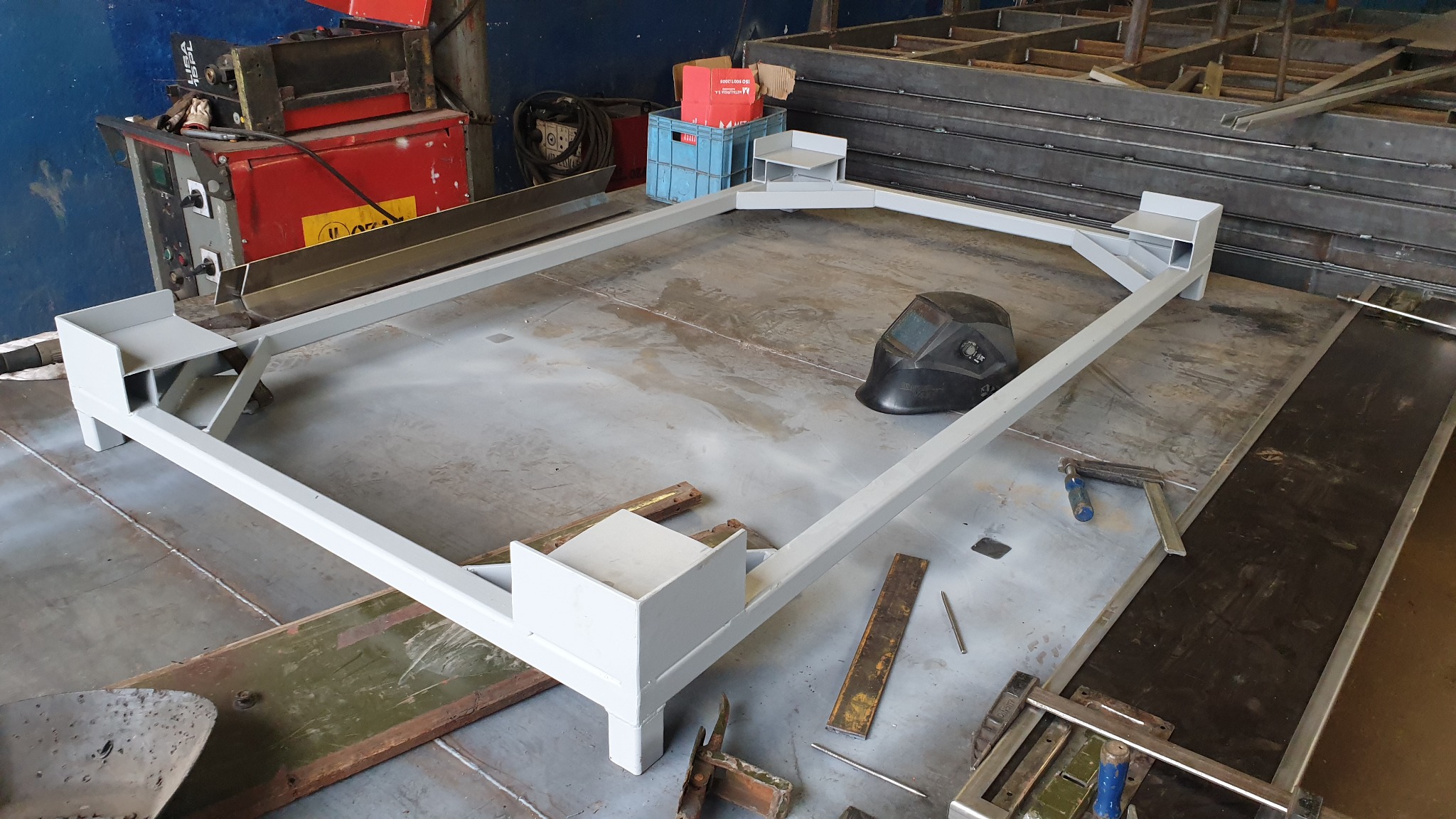 Stalowa rama konstrukcyjna w trakcie budowy, pomalowana na szaro, widoczna maska spawalnicza i narzędzia w warsztacie.