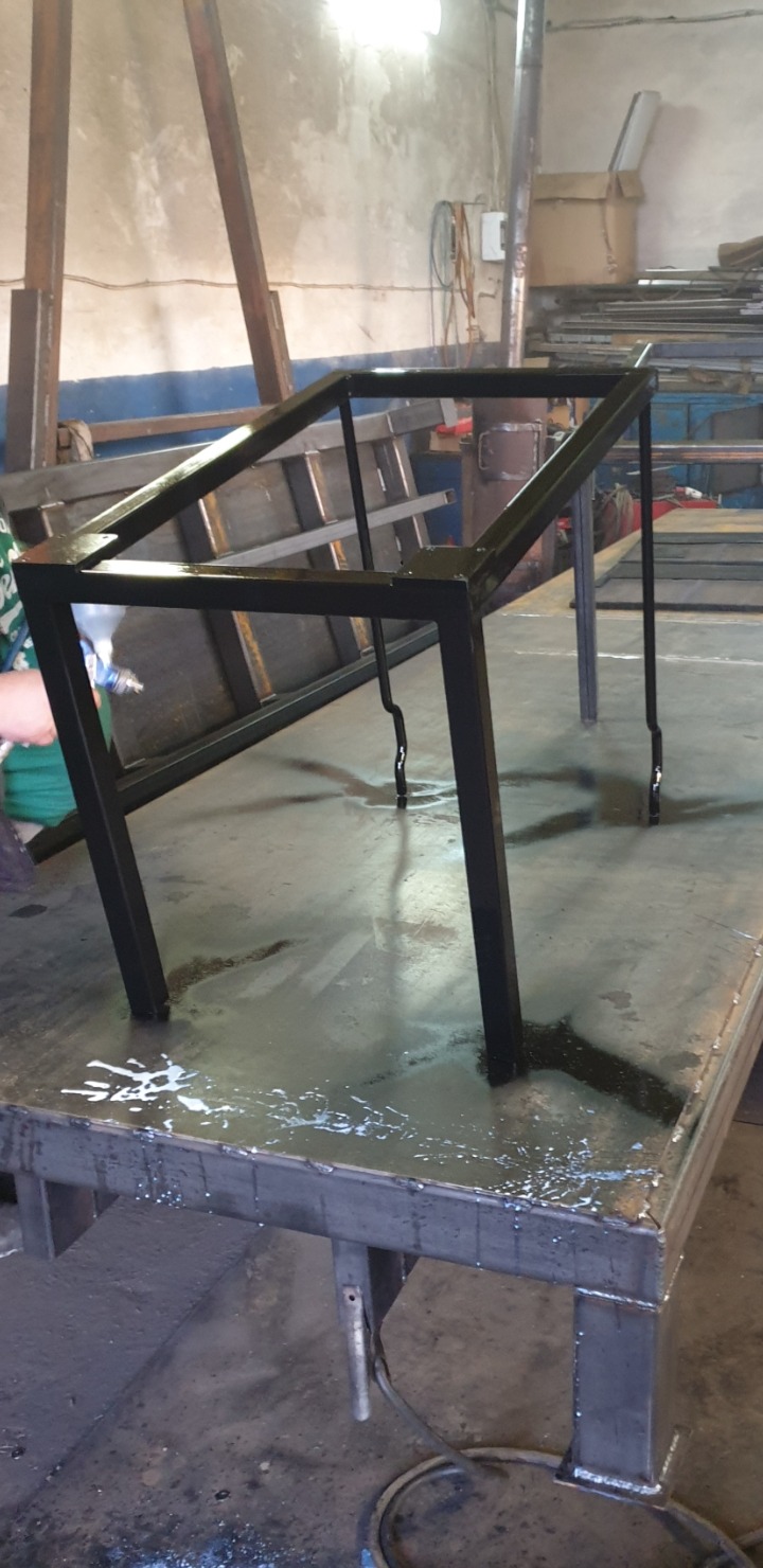 Malowanie natryskowe czarnej, metalowej konstrukcji stołu spawalniczego w warsztacie, z widocznymi śladami farby na stole roboczym.