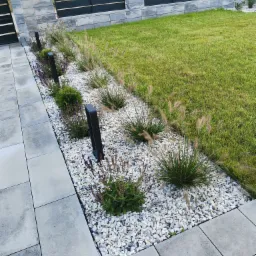 Nowoczesny ogród przydomowy z rabatą z ozdobnych traw i lawendy, oświetleniem punktowym oraz nawierzchnią z szarych płyt betonowych i białego grysu, ogrodzenie panelowe w tle.