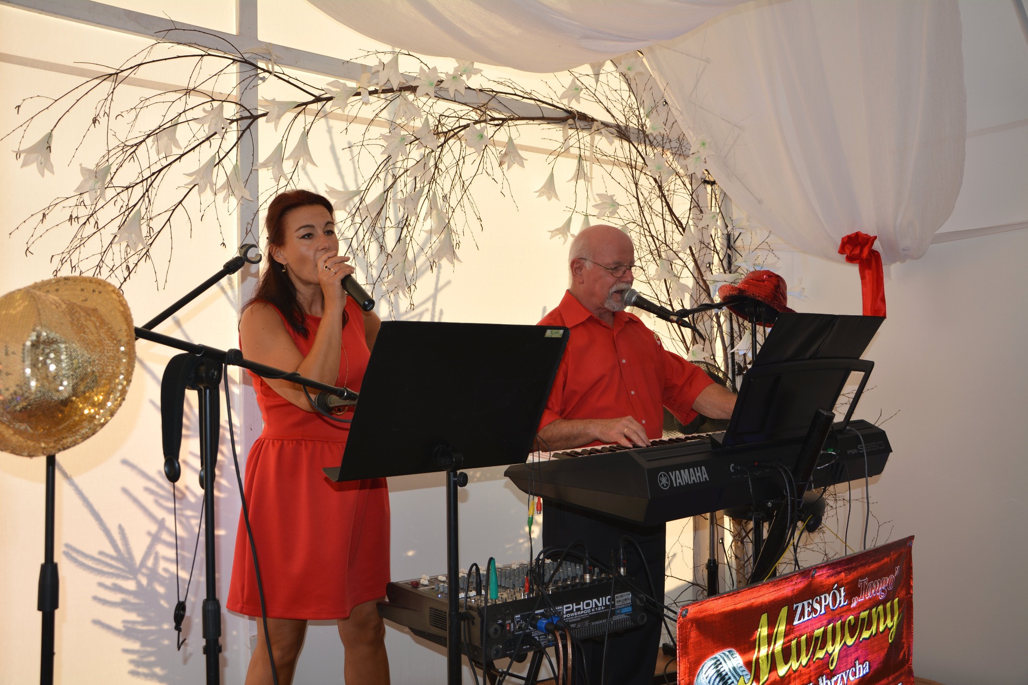 Wokalistka w czerwonej sukience śpiewa do mikrofonu obok klawiszowca grającego na keyboardzie Yamaha, scena udekorowana białymi kwiatami i tiulem, złoty kapelusz na statywie po lewej stronie.