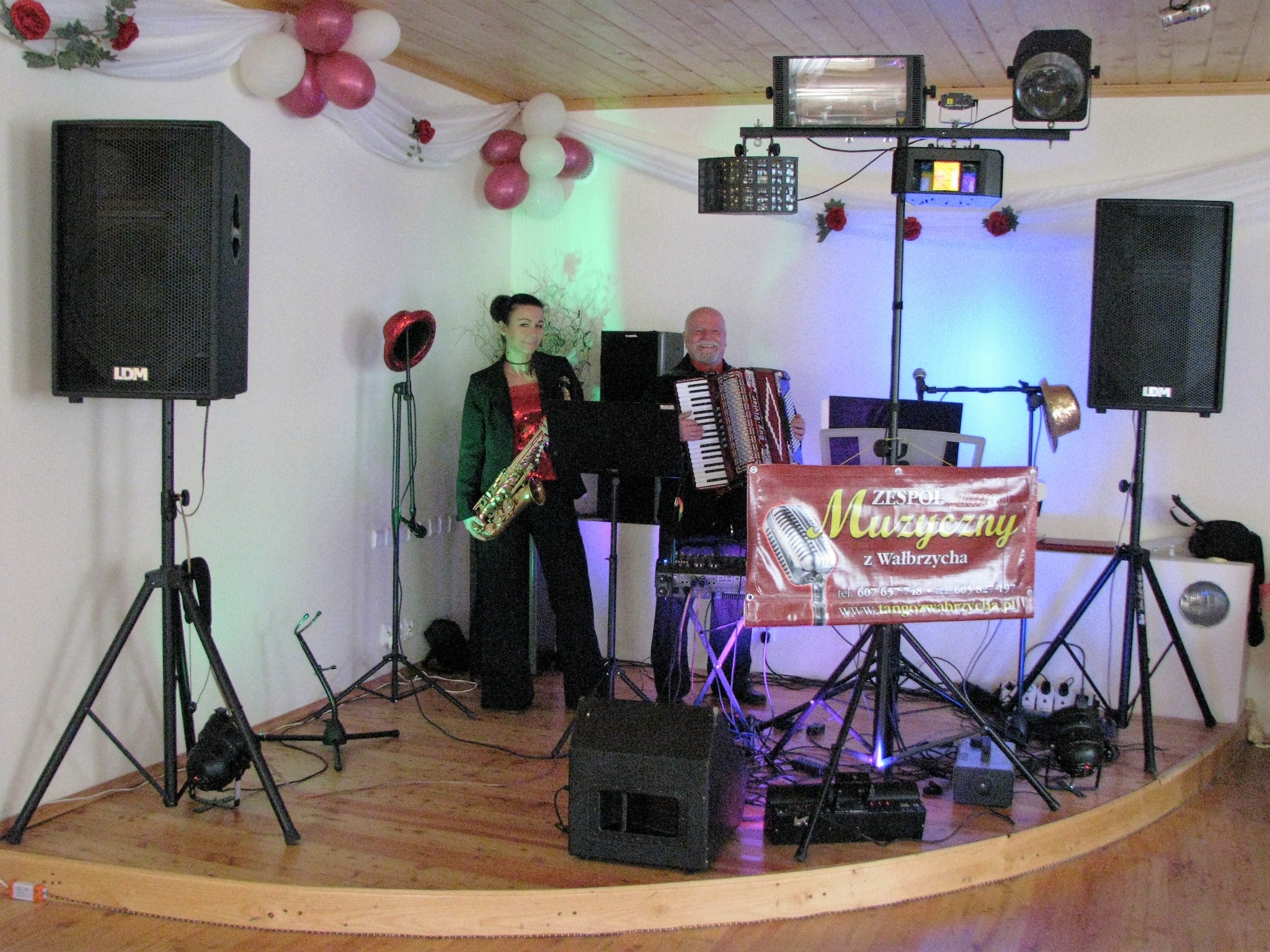 Duet muzyczny na scenie: kobieta z saksofonem i mężczyzna grający na akordeonie, nagłośnienie LDM, oświetlenie sceniczne, dekoracje z balonów i kwiatów.