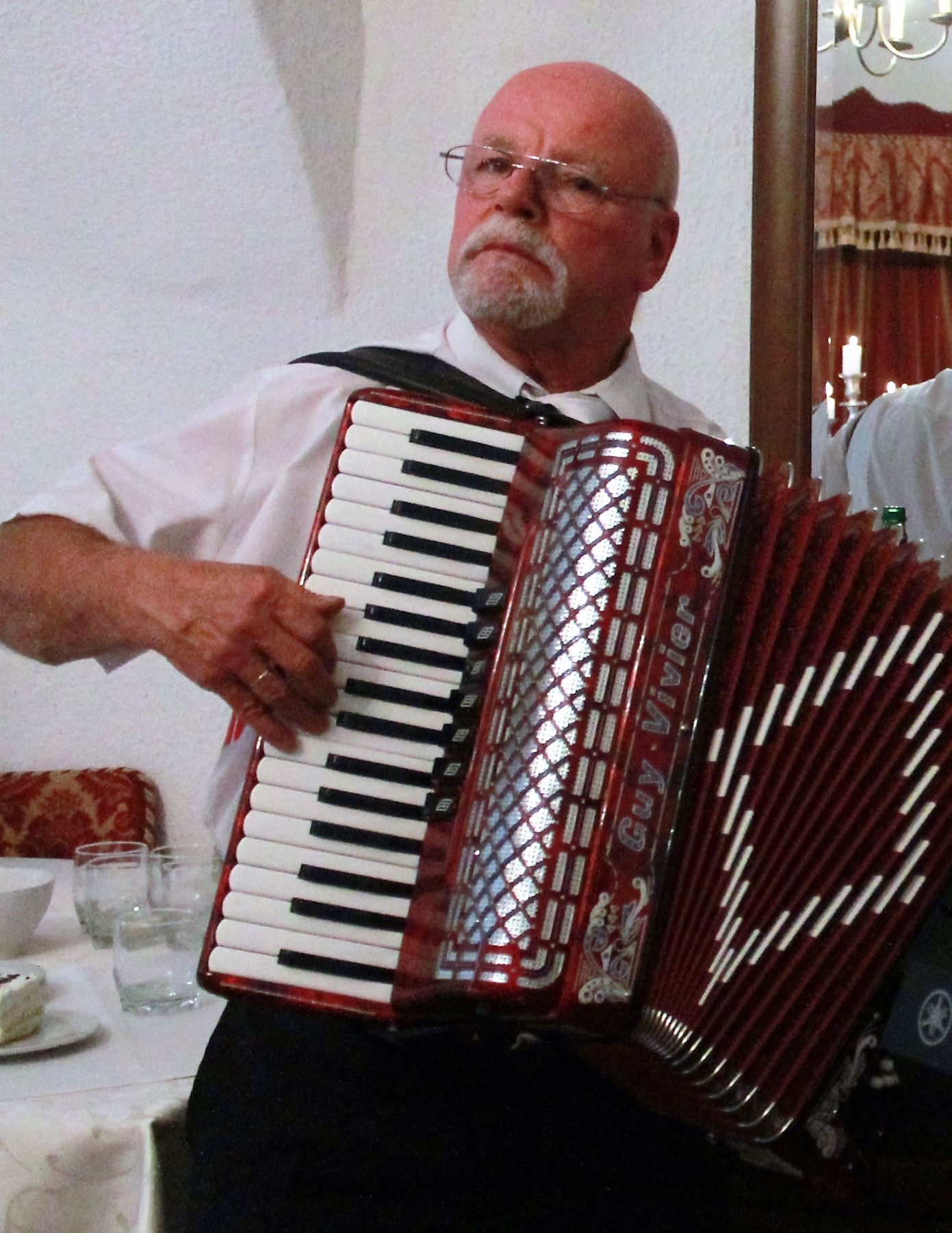Mężczyzna w okularach grający na czerwonym akordeonie marki Guy Vivier podczas uroczystości, w tle zastawiony stół i odbicie w lustrze.