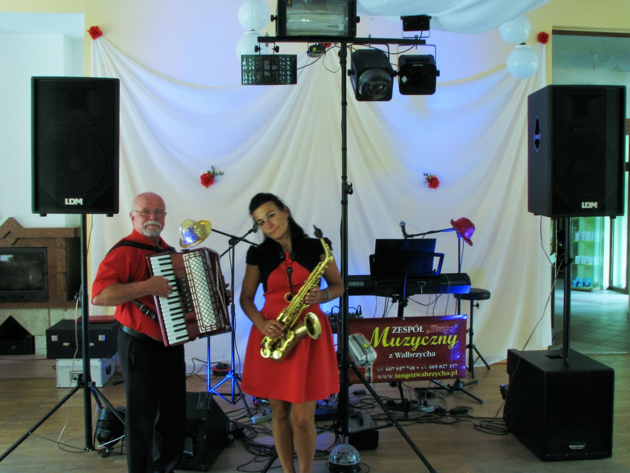 Duet muzyczny: mężczyzna z akordeonem i kobieta z saksofonem, na scenie z nagłośnieniem, oświetleniem i keyboardem, przed białym tłem z dekoracjami kwiatowymi.