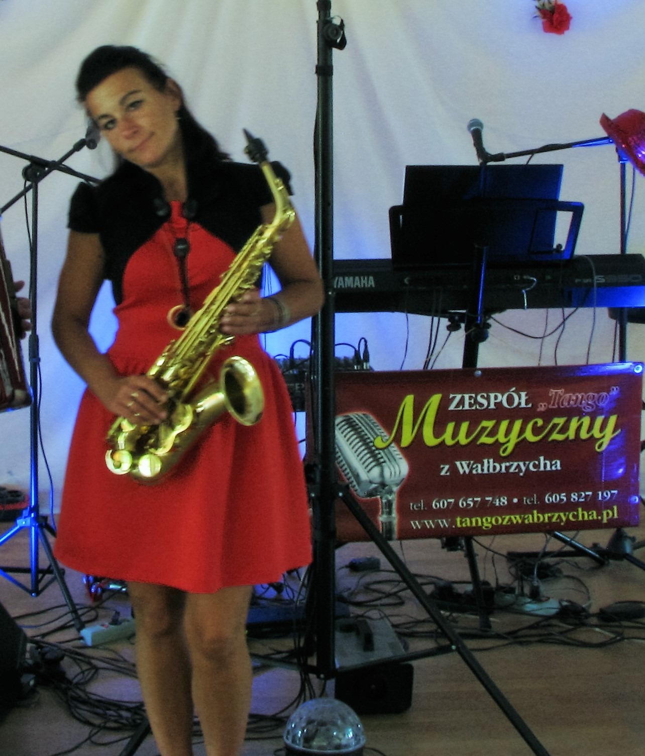 Uśmiechnięta kobieta w czerwonej sukience trzymająca saksofon na scenie z mikrofonami, keyboardem Yamaha i banerem reklamowym 'Zespół Tango Muzyczny z Wałbrzycha'.
