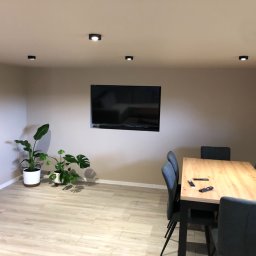 ITS PRO Tomasz Bar - Jasne wnętrze jadalni z drewnianym stołem i szarymi krzesłami, telewizor na ścianie, rośliny doniczkowe w rogu, parkiet imitujący drewno, oświetlenie punktowe na suficie.