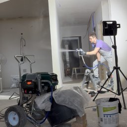 ITS PRO Tomasz Bar - Mężczyzna w okularach i kombinezonie maluje ściany wewnątrz budynku za pomocą profesjonalnego pistoletu natryskowego, z widocznym agregatem malarskim Groone i oświetleniem na statywie.