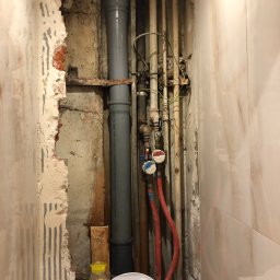 ITS PRO Tomasz Bar - Odkryte rury wodociągowe i kanalizacyjne w łazience podczas prac demontażowych, widoczne uszkodzenia ściany z resztkami kleju po płytkach, obok toaleta i słoik z kulkami dekoracyjnymi.