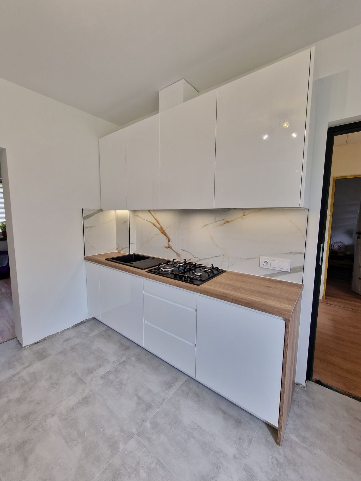 Nowoczesna kuchnia z białymi, błyszczącymi szafkami, blatem imitującym drewno i marmurowym panelem nad blatem, widok z boku na szarą podłogę z płytek.