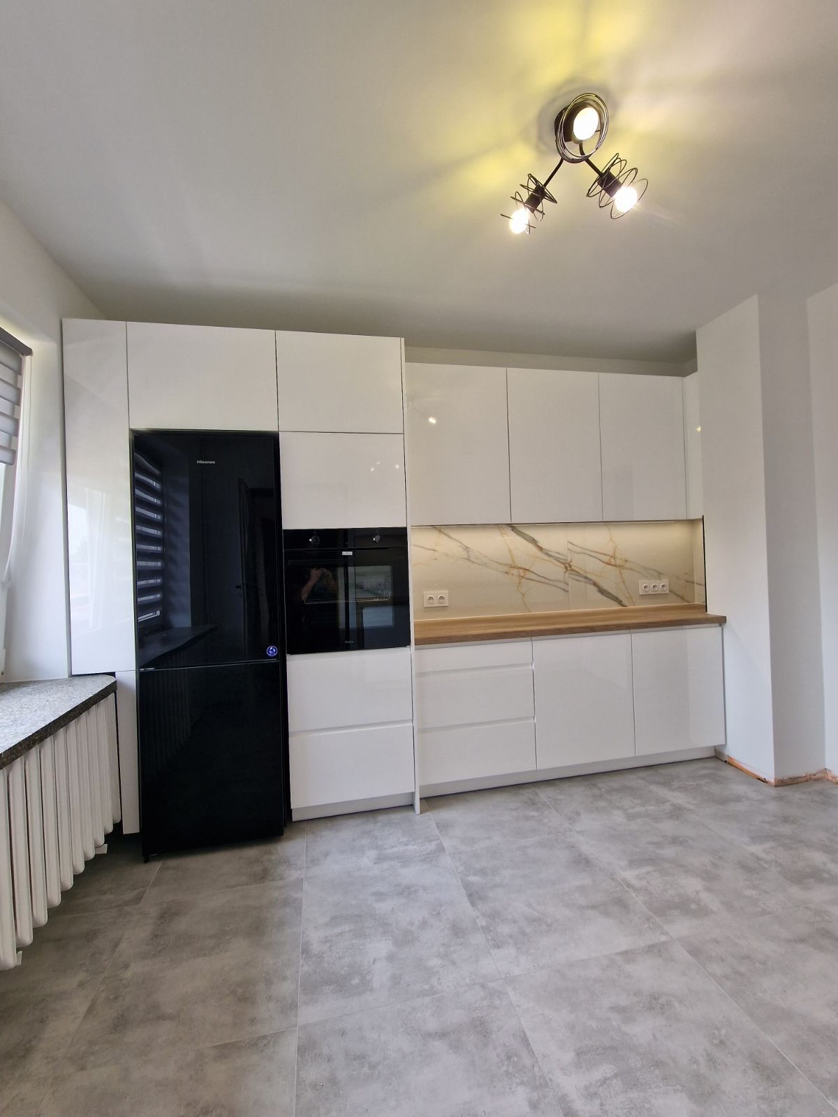 Nowoczesna kuchnia z białymi, błyszczącymi szafkami, czarną lodówką Hisense, marmurowym panelem nad blatem i szarą podłogą, oświetlona lampą sufitową z trzema żarówkami.