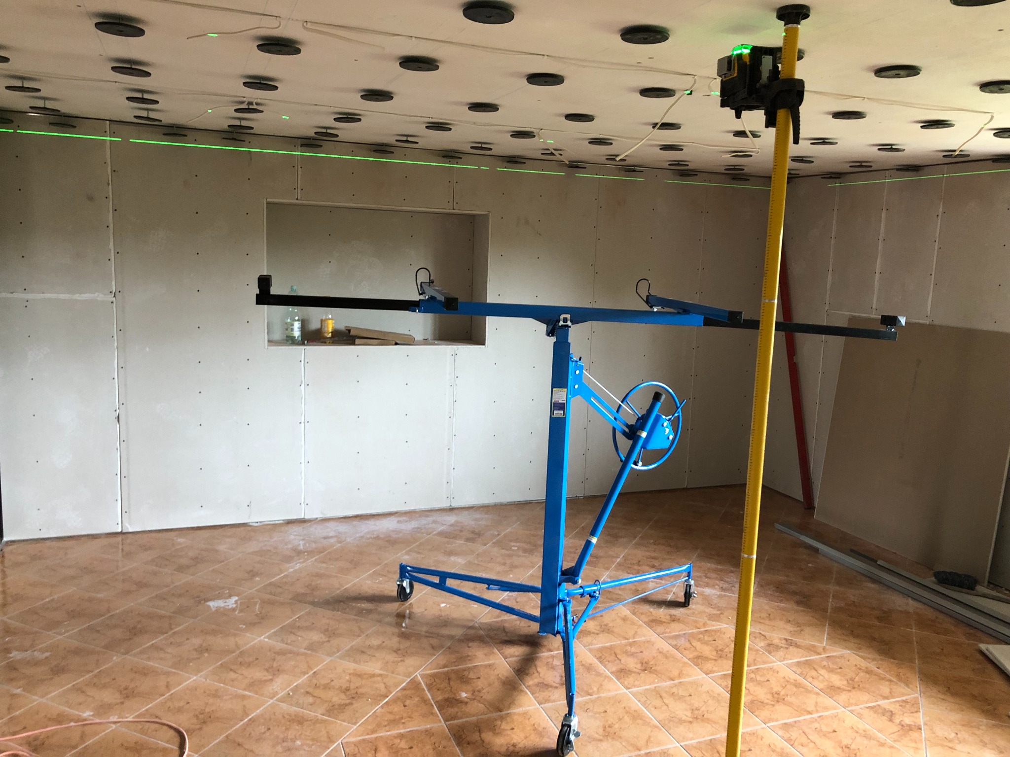 Montaż płyt gipsowo-kartonowych na suficie z użyciem podnośnika i lasera poziomującego, widoczne profile i elementy mocujące.