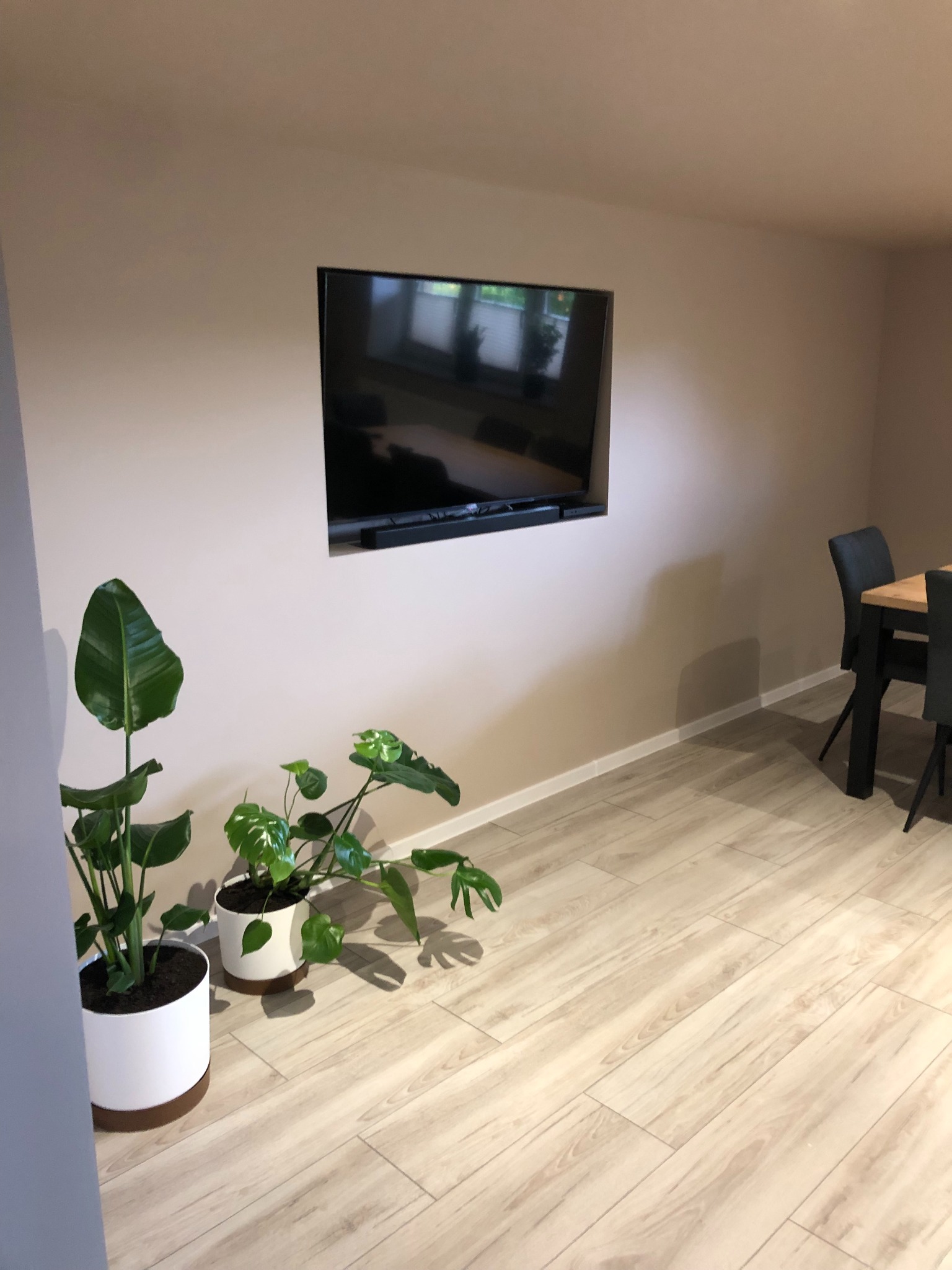 Telewizor wpuszczony w ścianę, pod nim soundbar, obok dwie rośliny doniczkowe, jasna podłoga imitująca drewno, stół z krzesłami w tle.