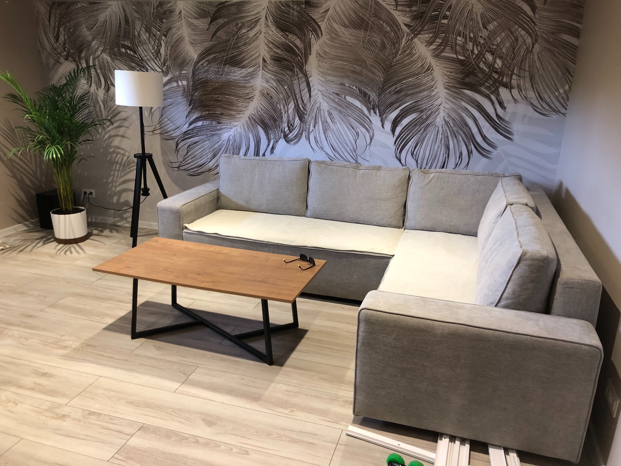 Jasnoszary narożnik z kremowymi poduchami, drewniany stolik kawowy z okularami, lampa stojąca i tapeta w liście w salonie z panelami podłogowymi.