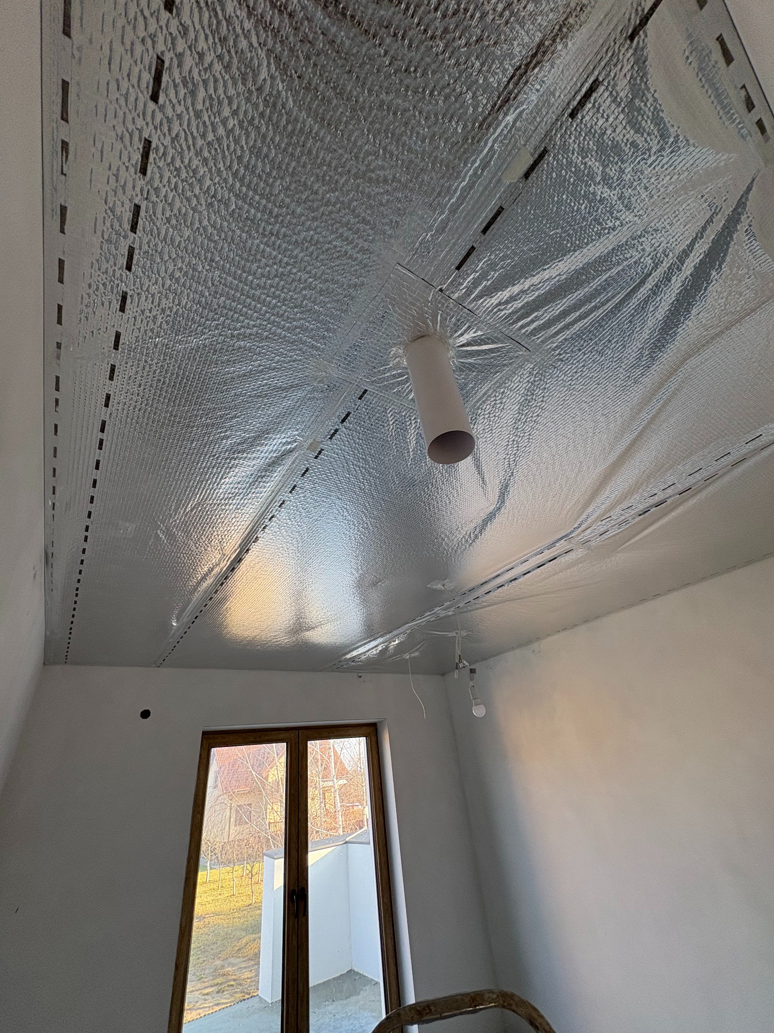 Montaż folii termoizolacyjnej na suficie poddasza, widoczne mocowania i otwór na oświetlenie, okno z widokiem na zewnątrz.