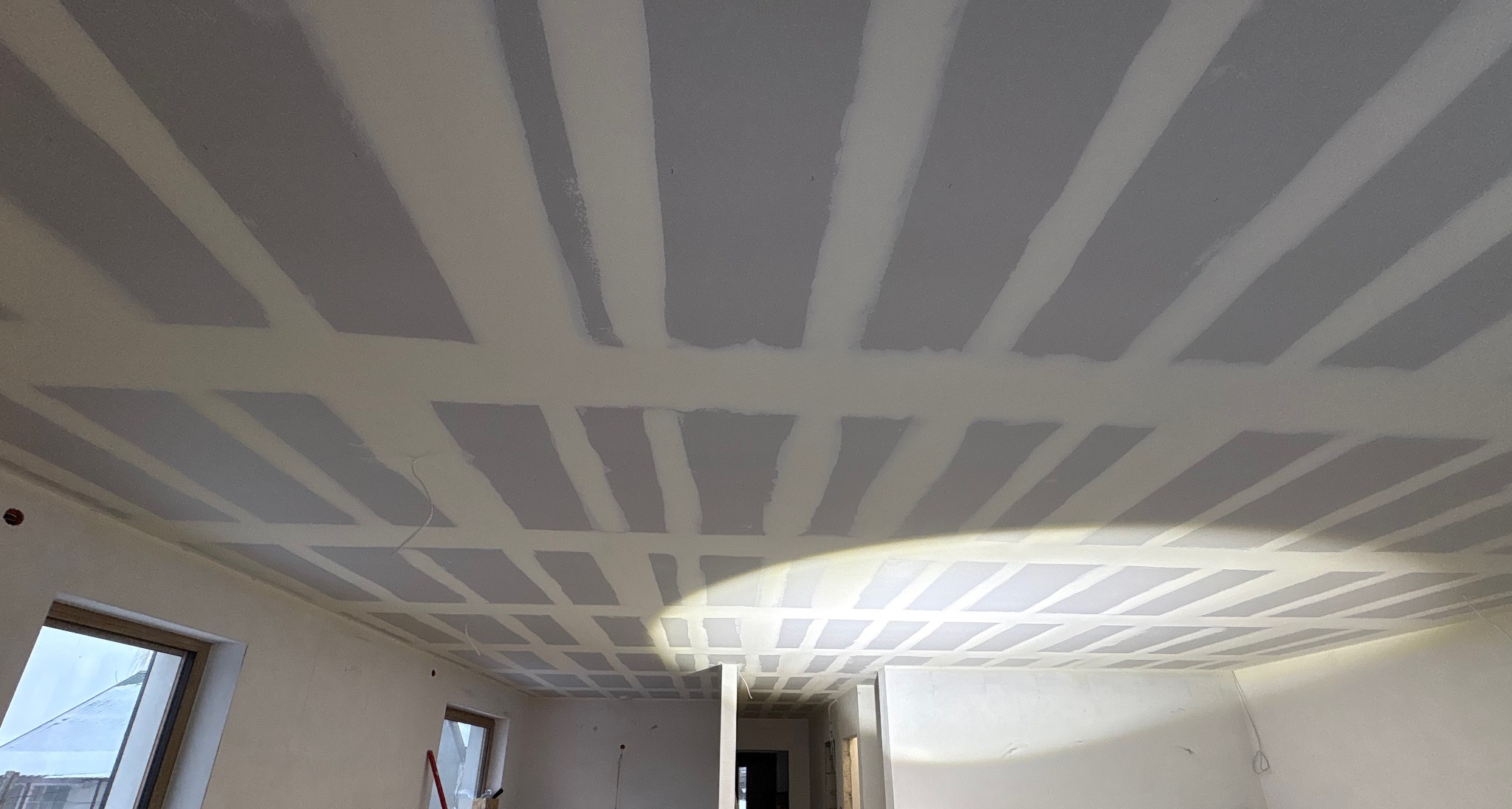 Sufit podwieszany z płyt gipsowo-kartonowych, szpachlowane łączenia, widoczne okna i fragment ściany w surowym stanie.