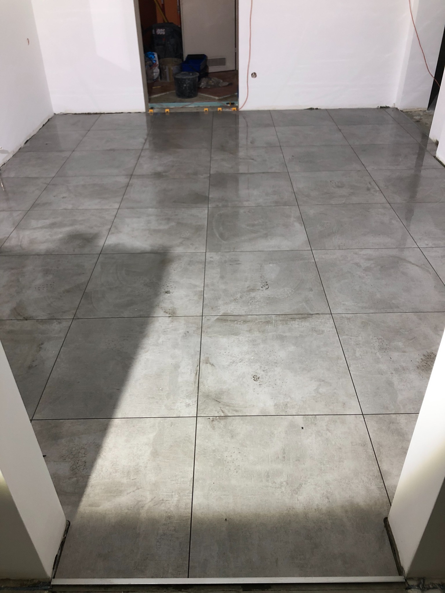 Świeżo położone, duże, kwadratowe, szare płytki podłogowe w trakcie schnięcia, widoczne białe ściany i fragmenty narzędzi w tle.