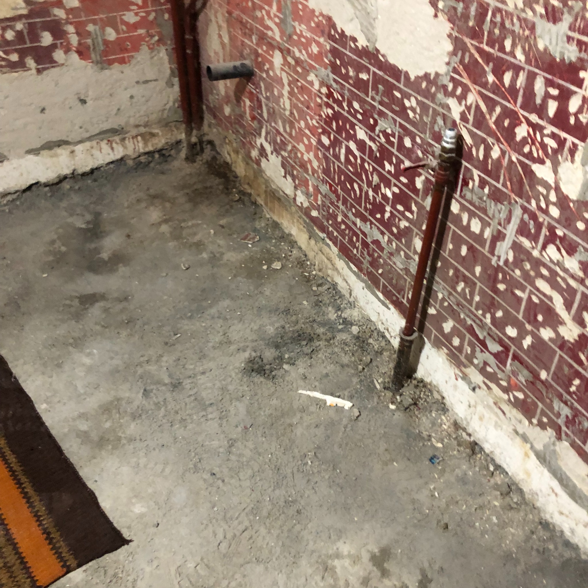 Pomieszczenie w trakcie demontażu wykończenia: fragment ściany z resztkami czerwonej glazury i odspojonym tynkiem, widoczne rury instalacji wodnej, surowy beton na podłodze z fragmentem dywanu w rogu.