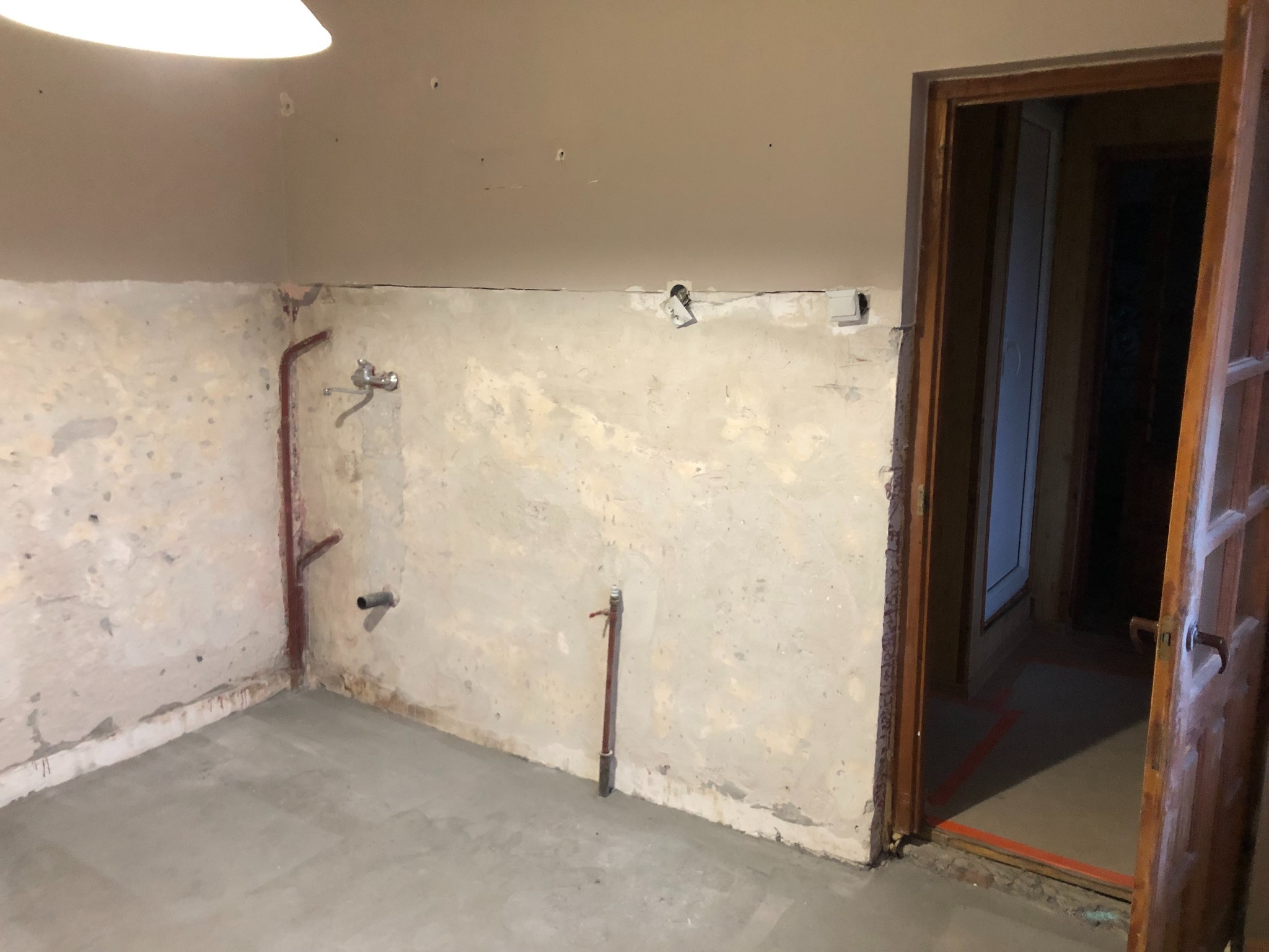 Wnętrze w trakcie prac przygotowawczych: surowe ściany z instalacjami, świeżo wylana podłoga, otwarte drewniane drzwi do kolejnego pomieszczenia.