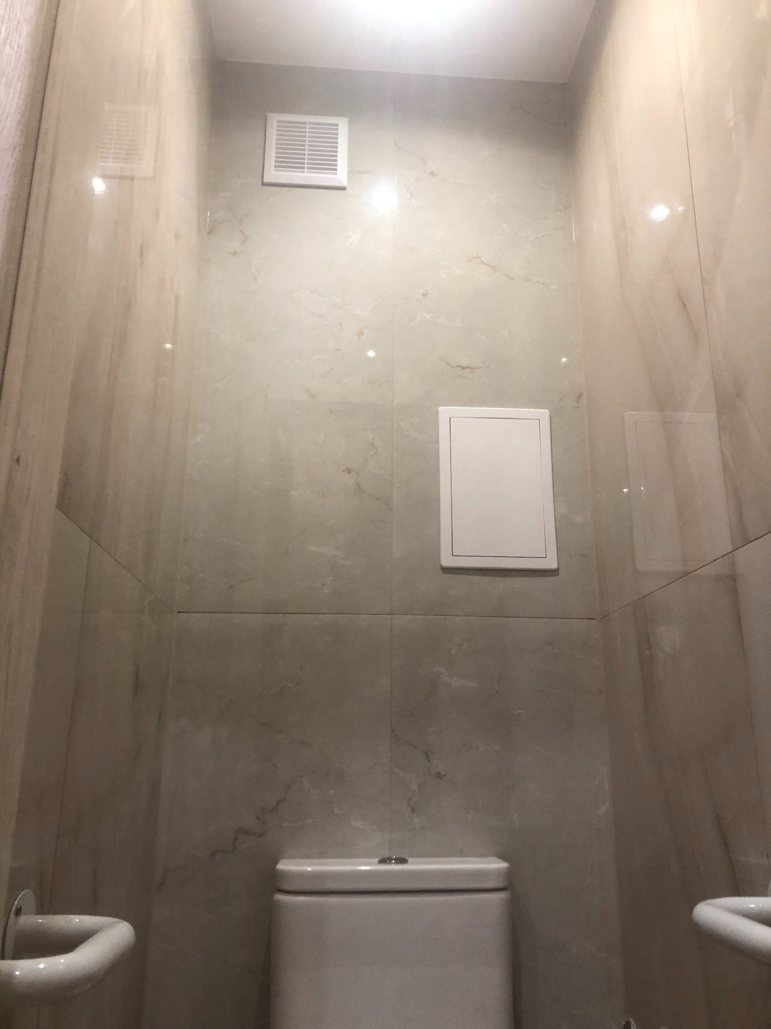 Wnętrze toalety wykończone płytkami imitującymi marmur, widoczna biała toaleta, wentylacja i rewizja na ścianie.