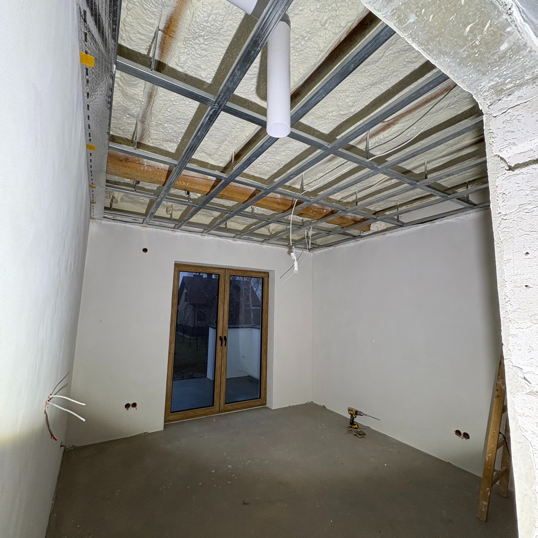 Wnętrze w trakcie prac wykończeniowych: sufit z metalową konstrukcją i pianą izolacyjną, ściany po gładziowaniu, widoczne przewody elektryczne, okno balkonowe i drabina w rogu.
