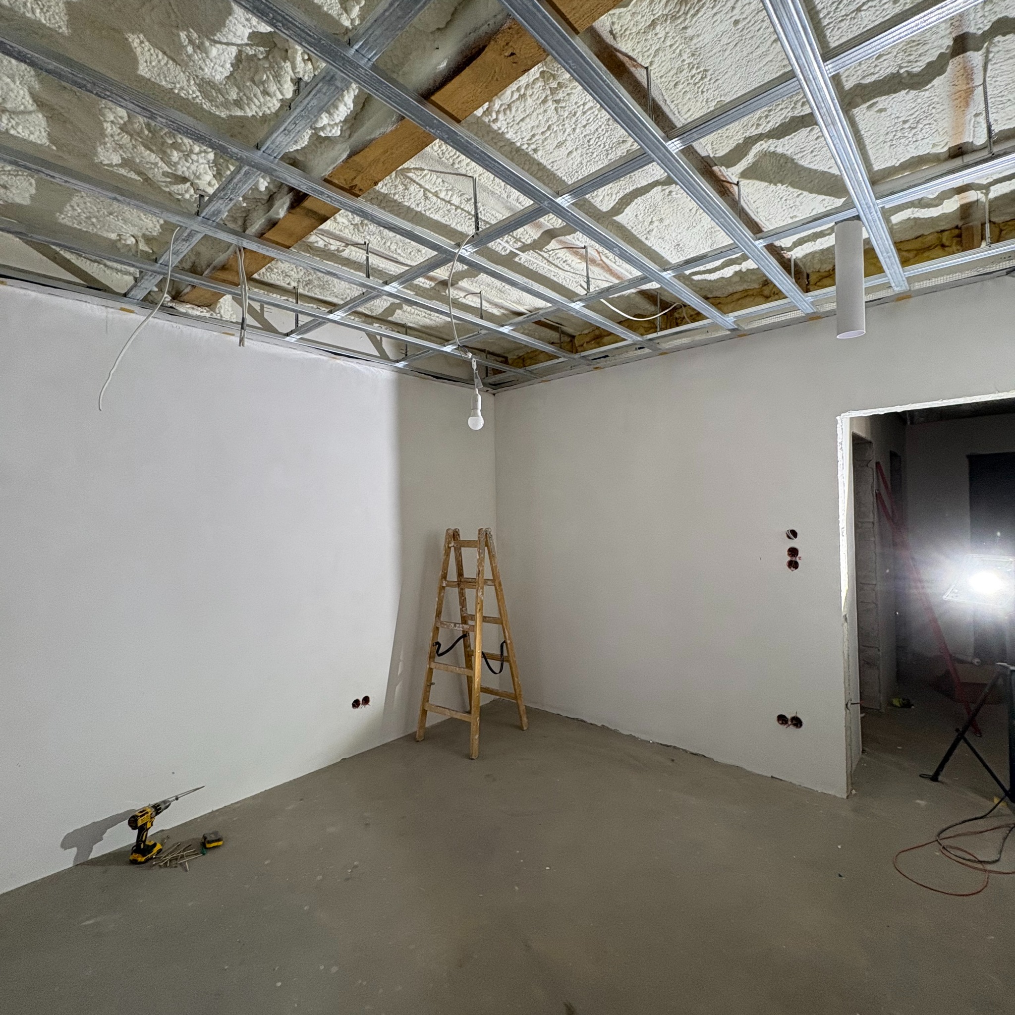 Wnętrze w trakcie prac: ocieplony pianą sufit z metalową konstrukcją podwieszaną, drabina drewniana, wiertarka z akcesoriami na szarym betonie, ściany przygotowane pod malowanie z widocznymi...
