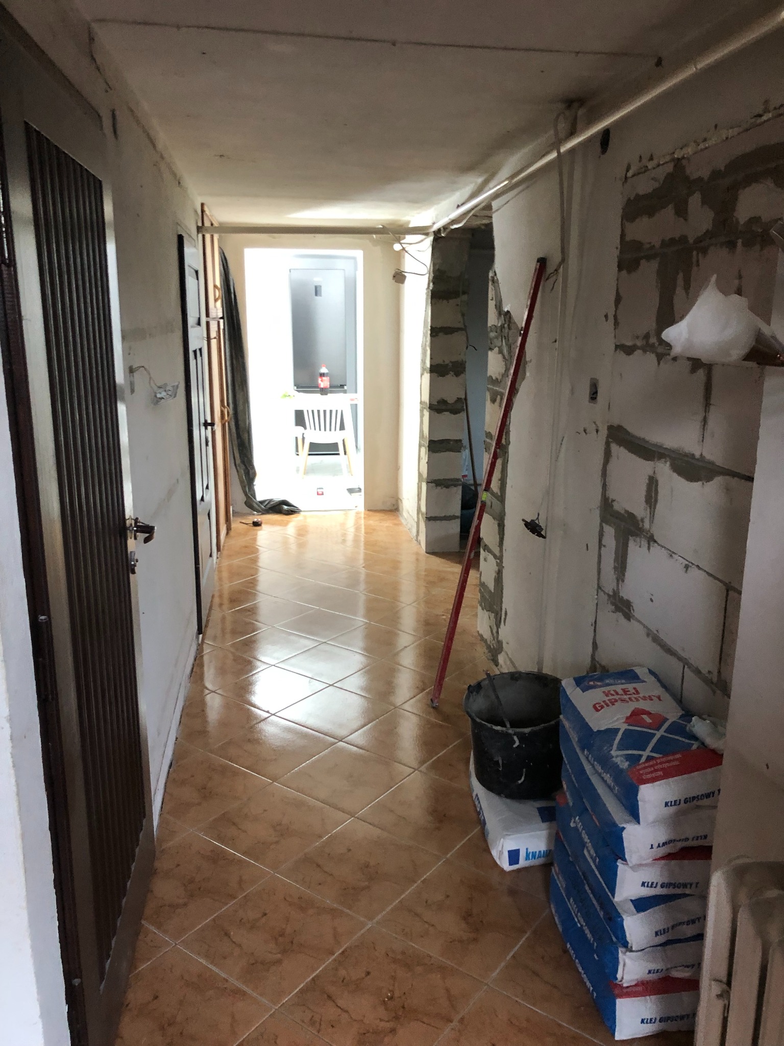 Korytarz w trakcie remontu: widoczne worki z klejem gipsowym, wiadro, poziomica oparta o ścianę, ściany w surowym stanie przed wykończeniem, brązowe płytki podłogowe.