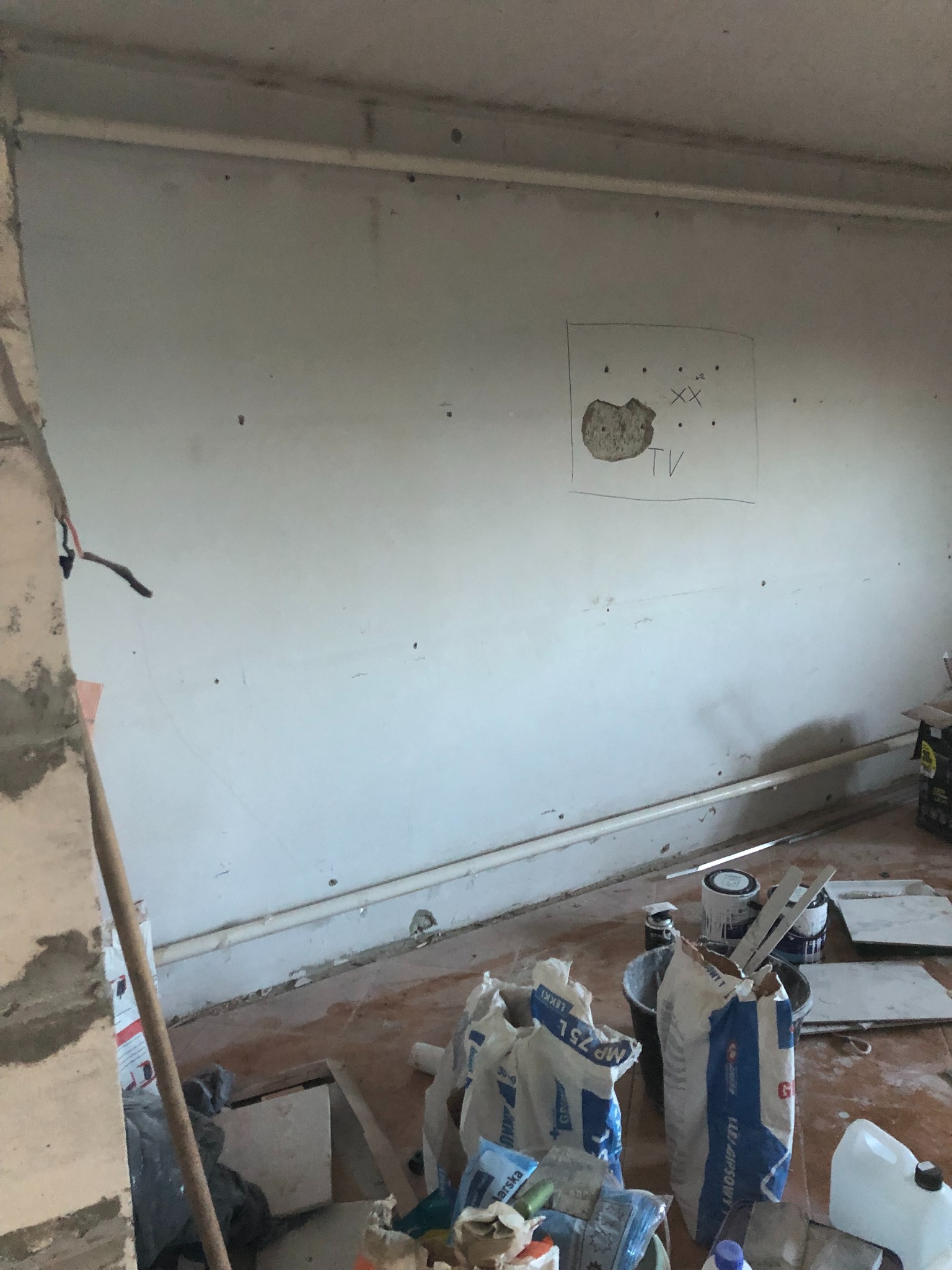 Wnętrze w trakcie prac wykończeniowych: surowa ściana z zaznaczonym miejscem na telewizor, worki z zaprawą gipsową i narzędzia malarskie na brudnej podłodze.