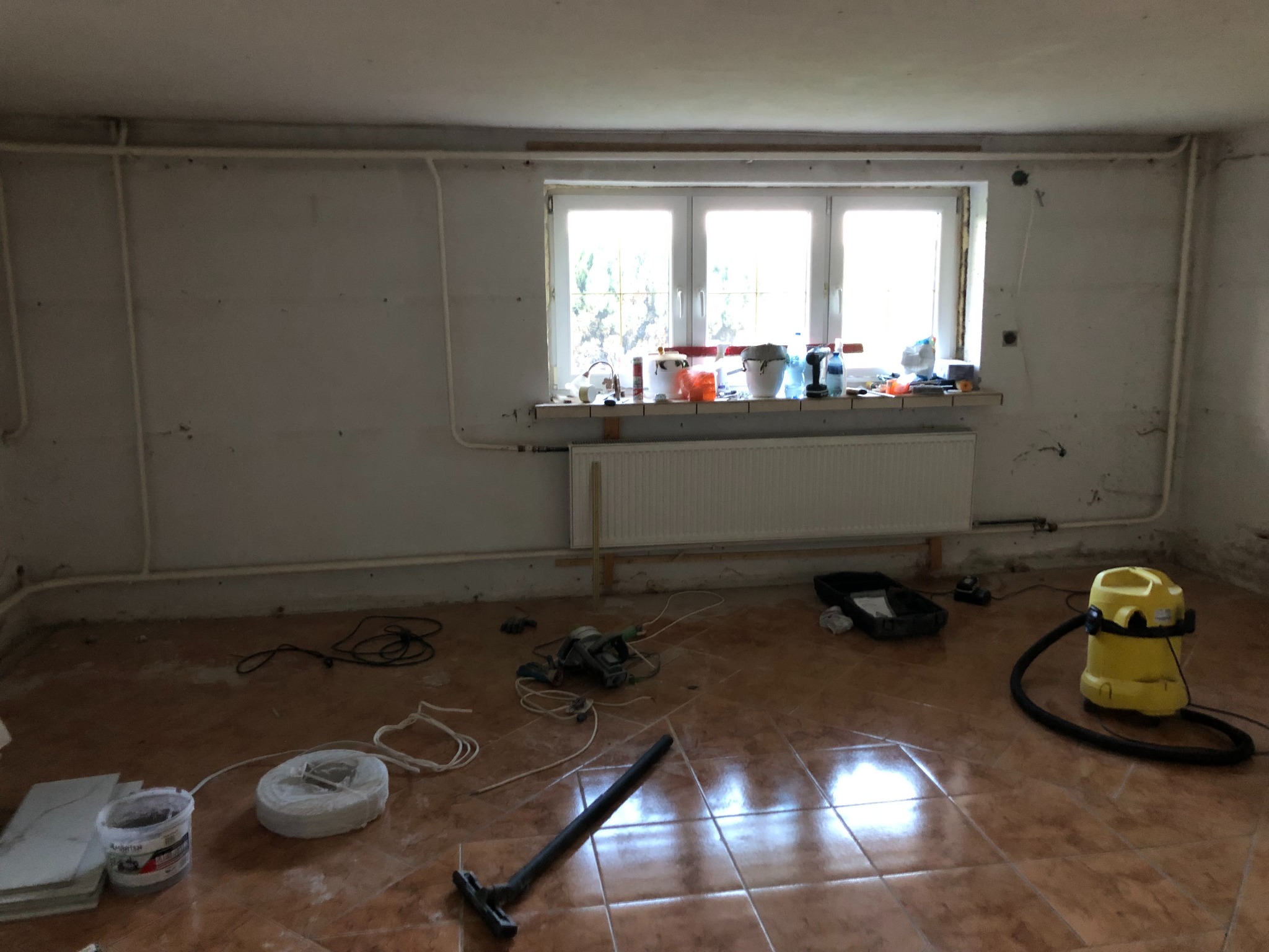 Pokój w trakcie prac przygotowawczych do remontu: widoczne narzędzia, wiadro z farbą, rolka folii malarskiej, odkurzacz przemysłowy, grzejnik pod oknem, ściany z widocznymi śladami po...