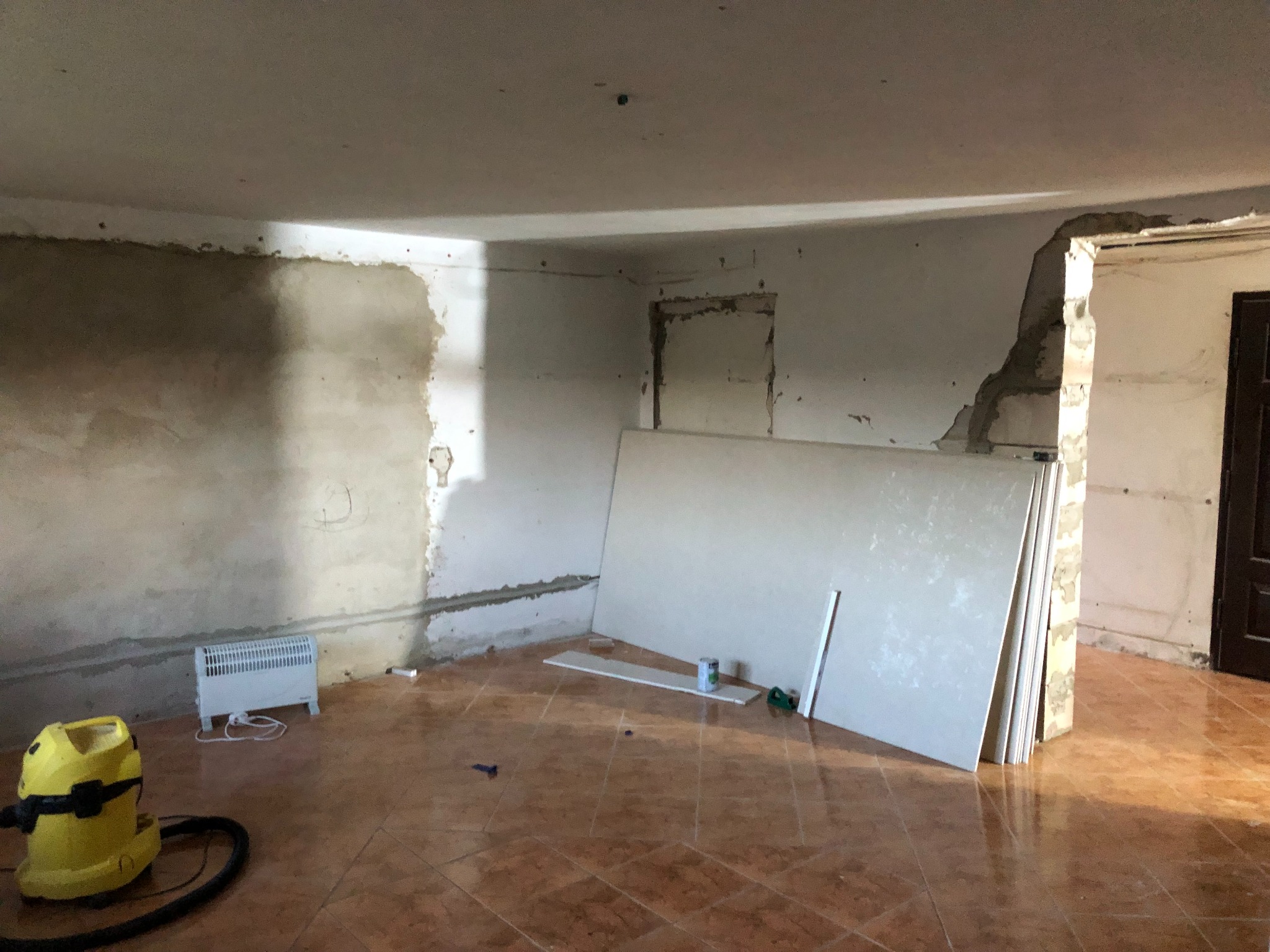 Pokój w trakcie prac wykończeniowych: ściany częściowo otynkowane, płyty gipsowo-kartonowe oparte o ścianę, grzejnik elektryczny i żółty odkurzacz przemysłowy na brązowej podłodze z płytek.