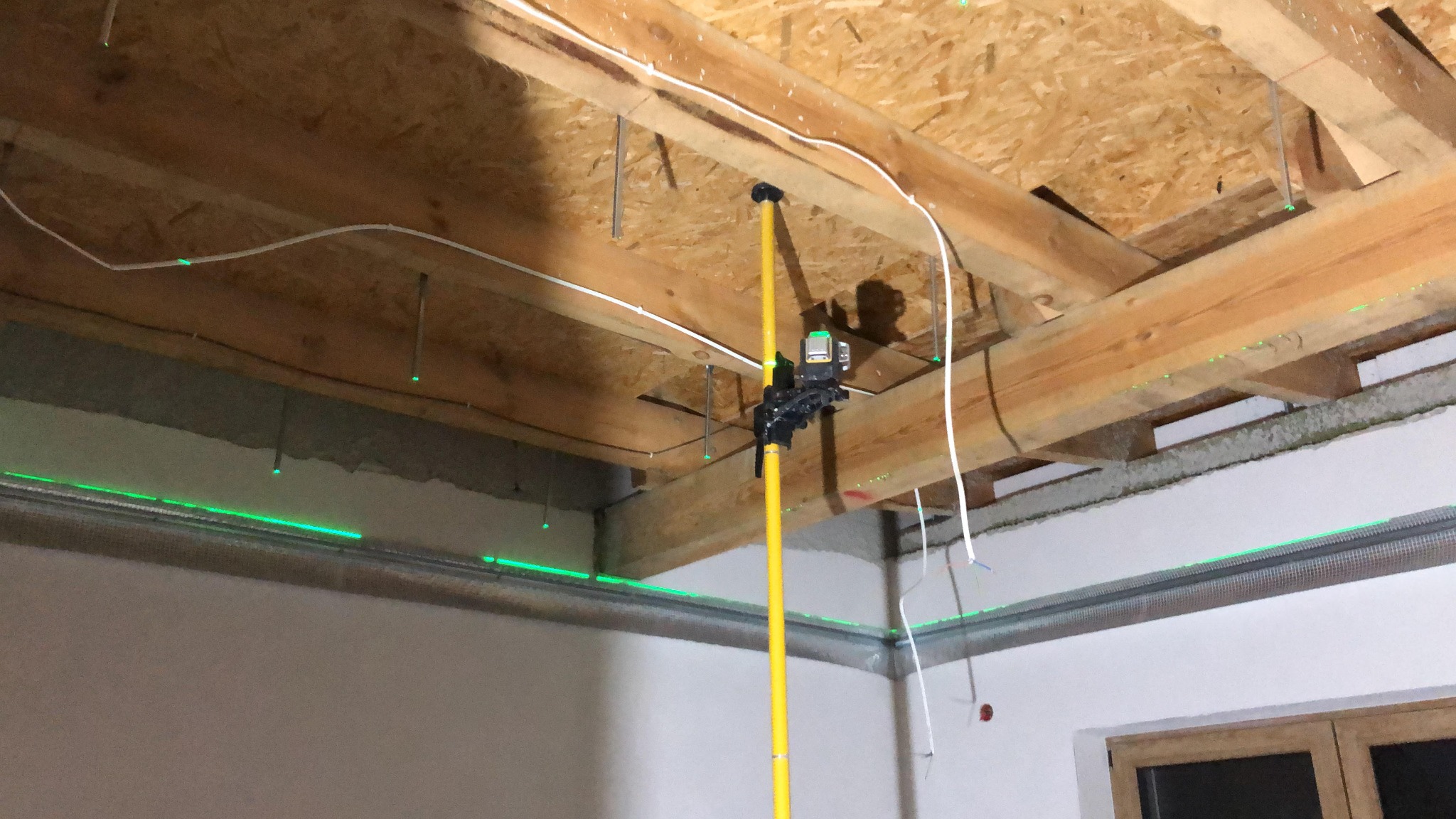 Laser krzyżowy na tyczce wyznacza poziom na ścianach pomieszczenia z widoczną konstrukcją drewnianą sufitu podwieszanego i przewodami elektrycznymi.