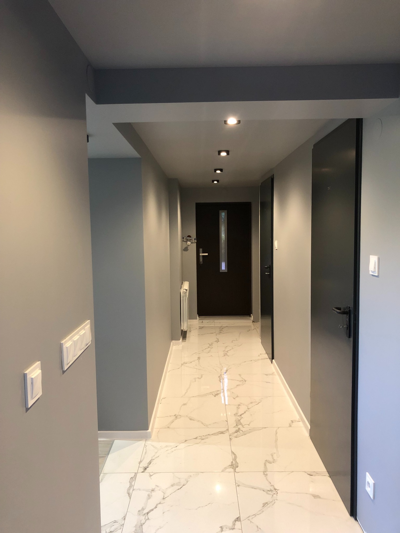 Korytarz po odświeżeniu: szare ściany, marmurowe płytki podłogowe, ciemne drzwi i minimalistyczne oświetlenie sufitowe