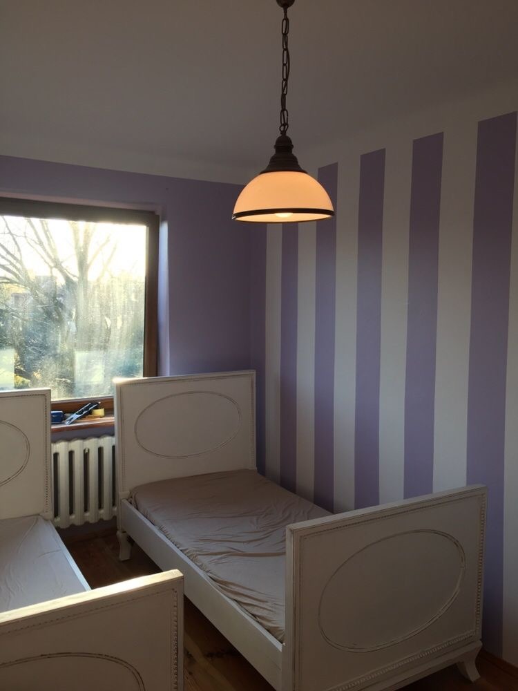 Pokój dziecięcy z dwoma białymi łóżkami, ściany w pionowe biało-lawendowe pasy, wisząca lampa z białym kloszem, okno z widokiem na drzewa.