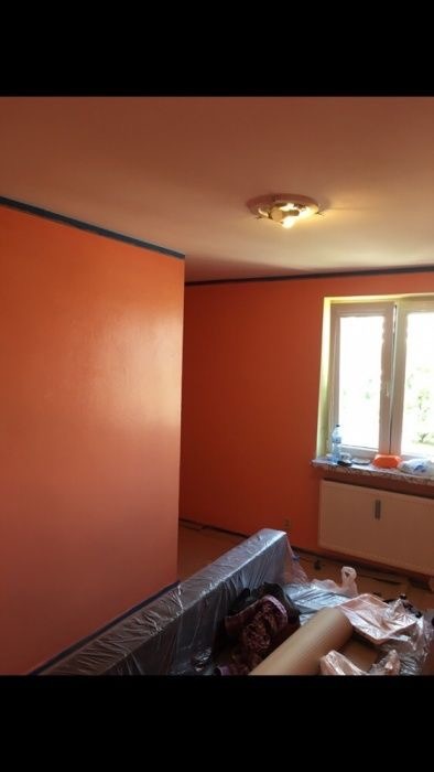 Pomarańczowe ściany w pokoju po świeżym malowaniu, zabezpieczone listwy przypodłogowe i okno, meble zakryte folią ochronną.